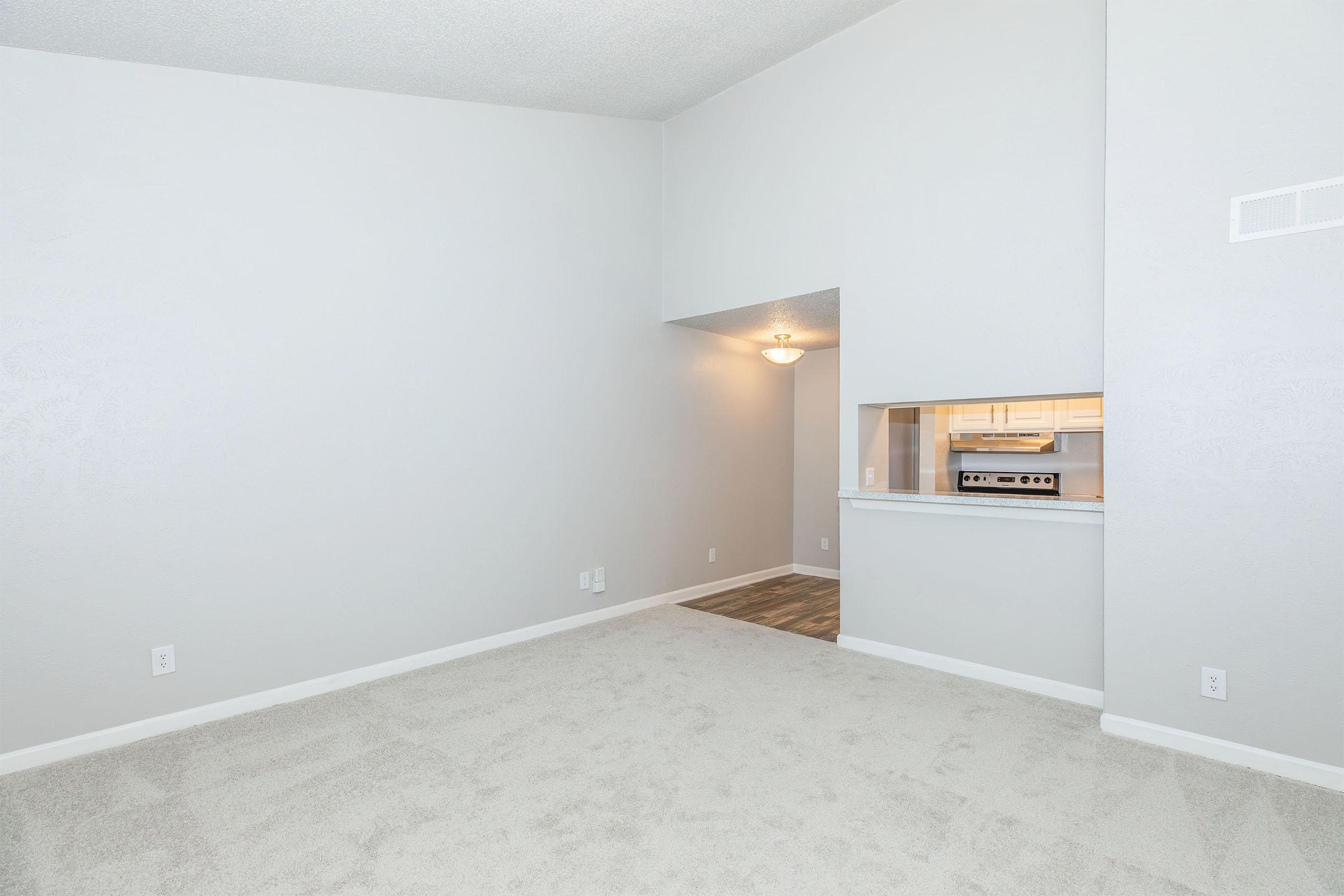 Bright, spacious living area with light gray walls and plush carpeting. The room features a high ceiling and an open layout leading to a kitchen area, partially visible through an archway. Soft lighting adds warmth to the space, creating a welcoming atmosphere.