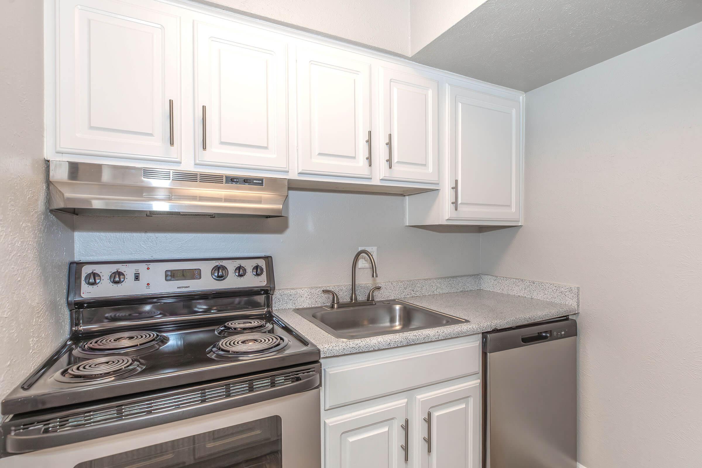 A modern kitchen featuring white cabinetry, a stainless steel stove with an oven, a sink with a chrome faucet, and a silver dishwasher. The countertops are grey with a subtle pattern, and the walls are painted in a neutral color, creating a clean and contemporary look.