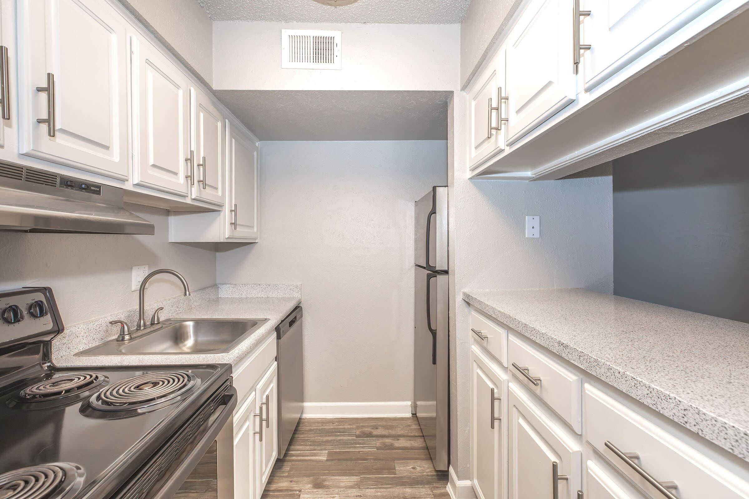 A modern kitchen featuring white cabinetry, a stainless steel sink, and a black stove. The countertop is made of light-colored granite, and there is a refrigerator visible in the corner. The overall color scheme is neutral, with gray walls and wood-like flooring, creating a clean and inviting space.