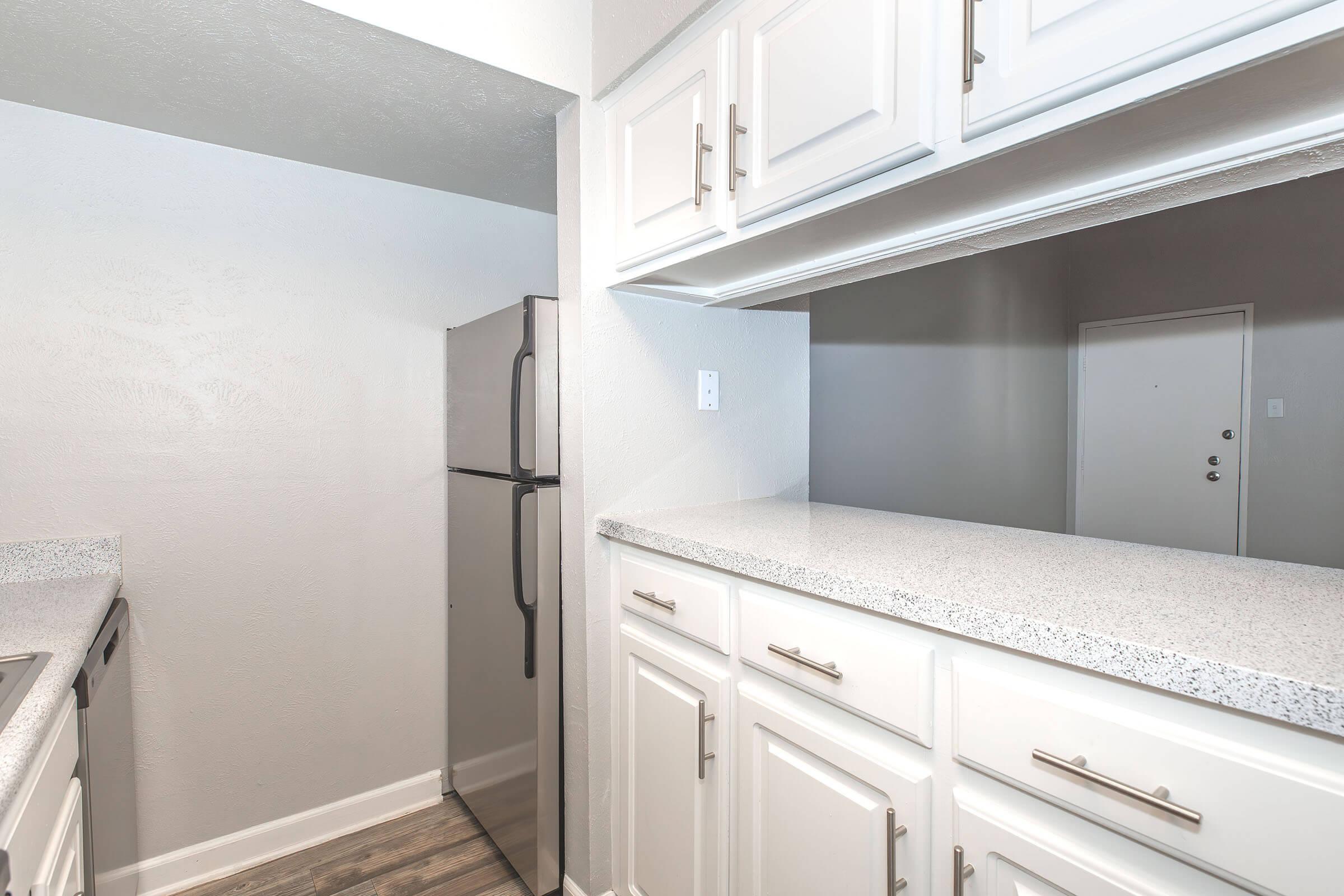 A modern kitchen featuring white cabinets, a countertop, and a stainless steel refrigerator. The space is well-lit with a neutral color scheme and an opening that connects to another room beyond.