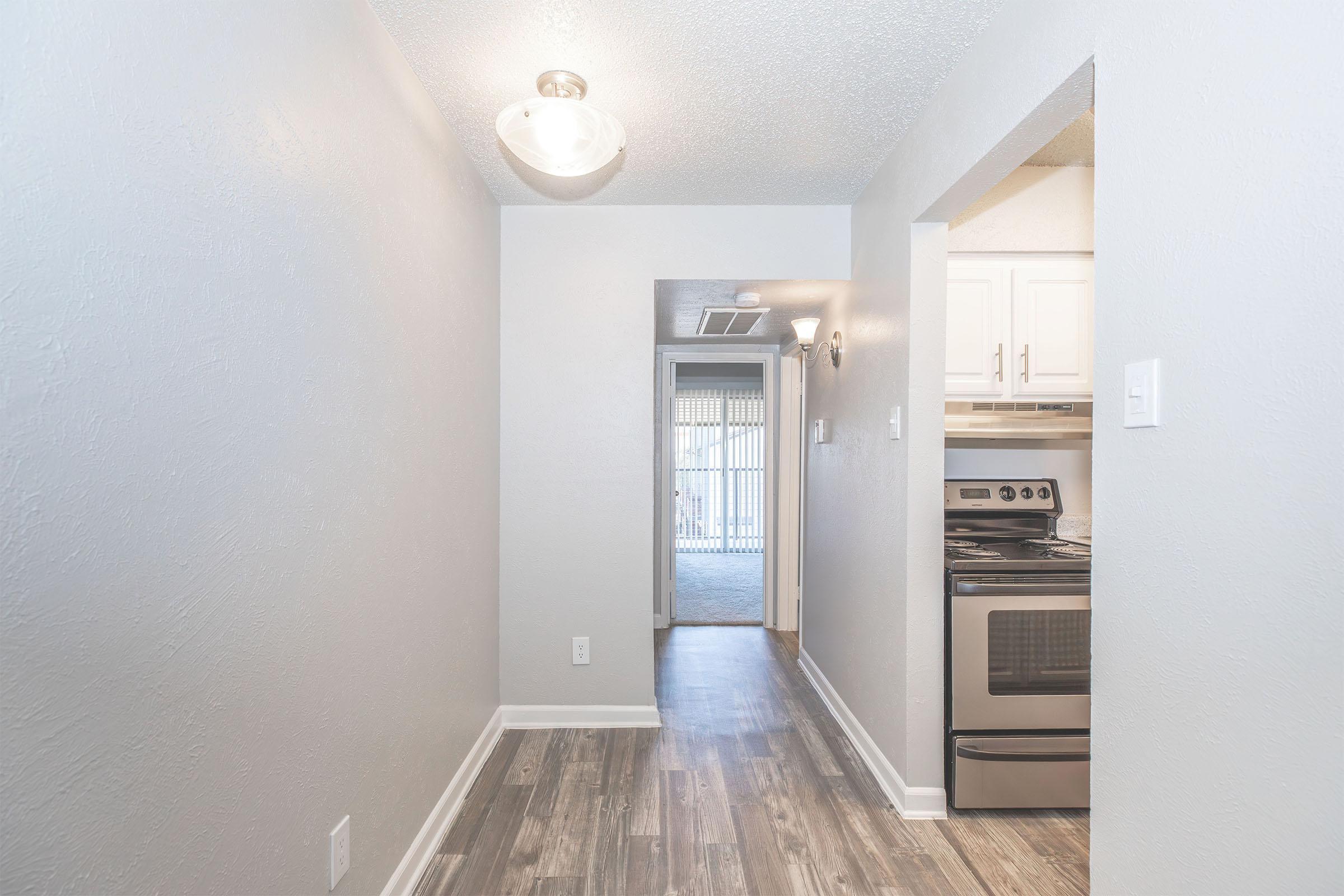 A well-lit hallway with light gray walls and modern flooring, leading to an open kitchen area on the left. A stainless steel stove is visible in the kitchen, while an entrance to a patio or outdoor space can be seen at the end of the hallway. The overall design is clean and contemporary.