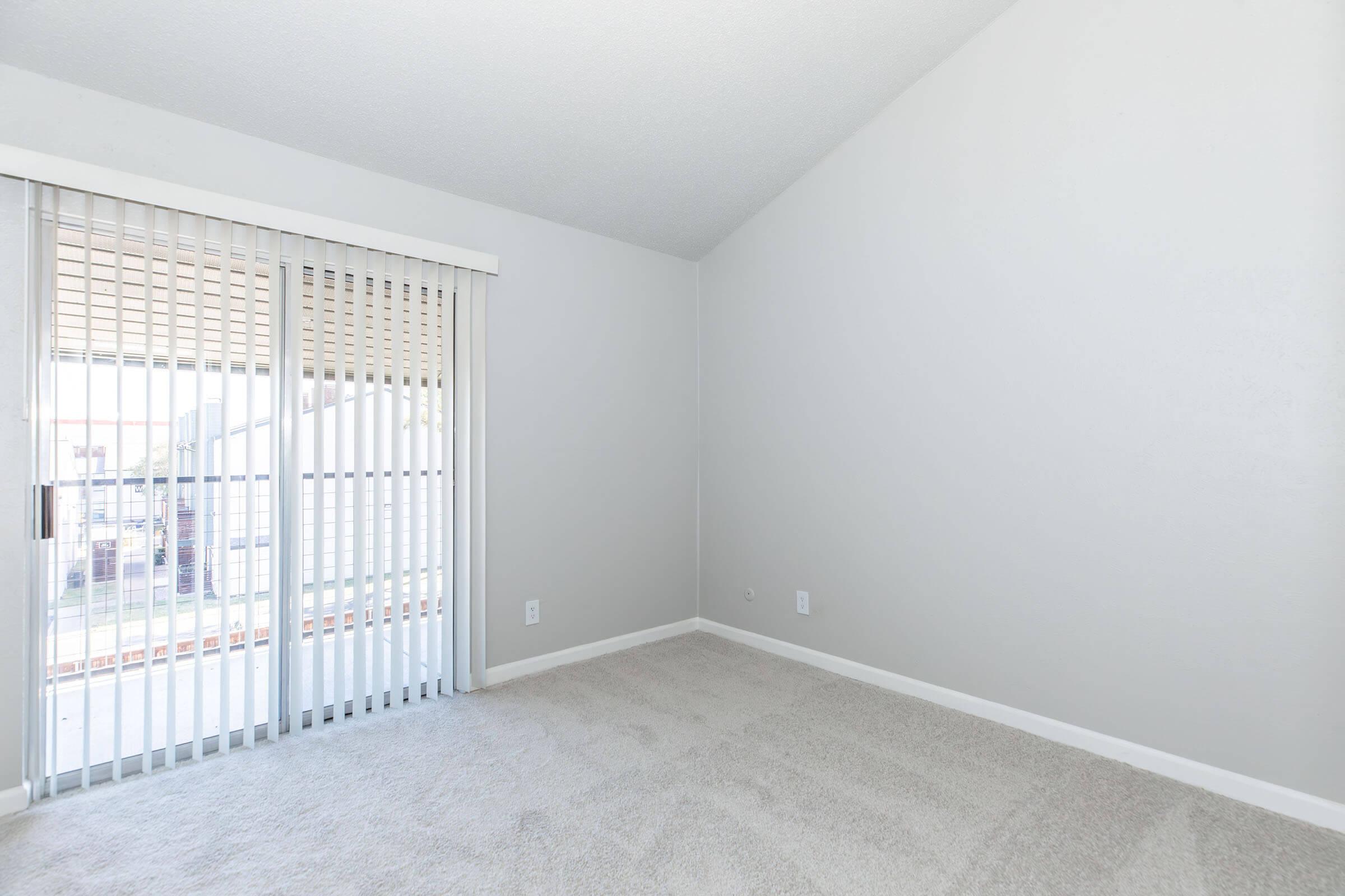 Empty room with light gray walls and light carpet. Features large sliding glass doors with vertical blinds that open to a patio or balcony. The ceiling is high, creating an airy feel. Natural light floods in through the door, making the space bright and inviting.