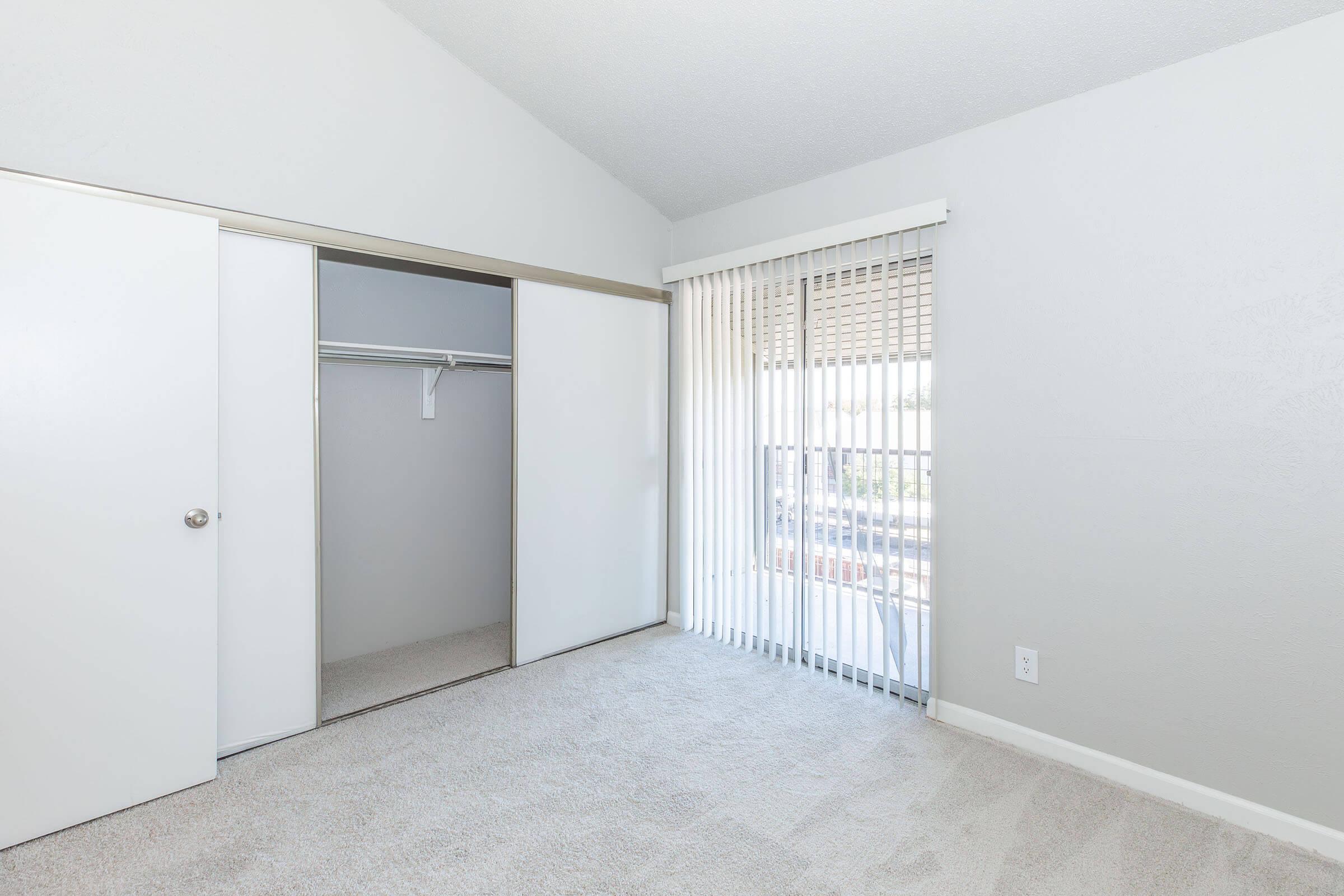 A bright, empty bedroom with light gray walls and carpet. It features a large window with vertical blinds, allowing natural light to enter, and a closet with sliding doors on one side. The room has a simple and clean aesthetic, suitable for personal decoration.