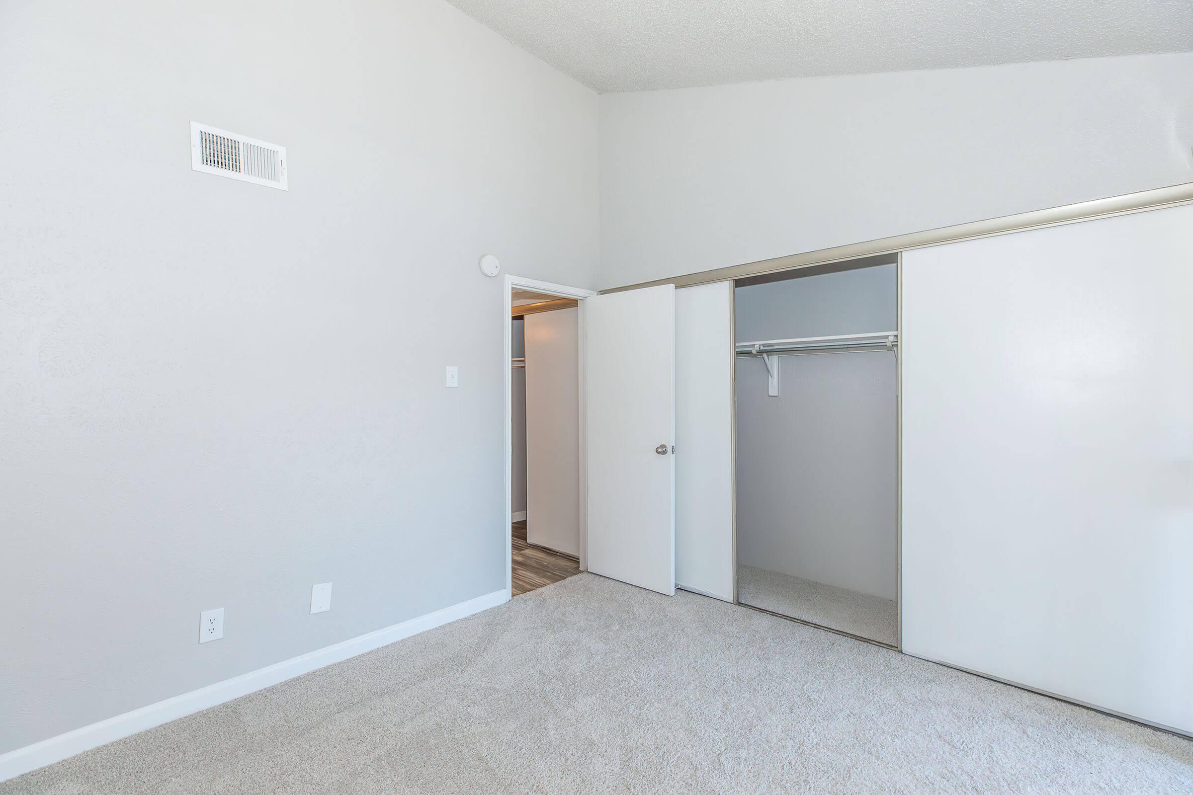 A room with light gray walls and carpet. There are two doors: one leads to a closet with a hanging rod, and the other opens into another space. The room has a high ceiling and no furniture, creating a spacious and airy feel.