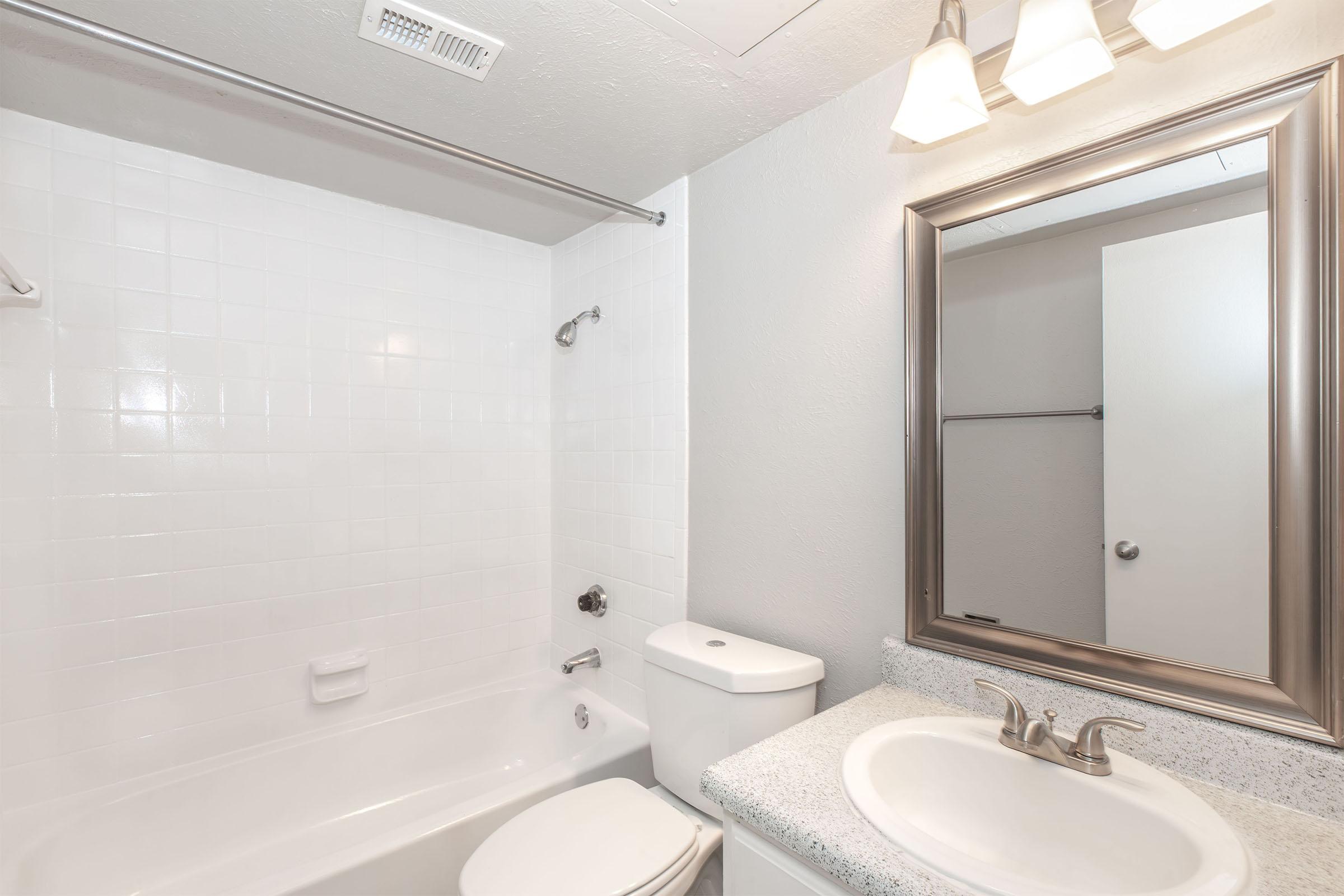 A bright, clean bathroom featuring a white tiled shower and bathtub combination. There is a modern sink with a silver faucet, a large framed mirror above it, and soft lighting from three light fixtures. The walls are painted a light color, adding to the spacious feel.