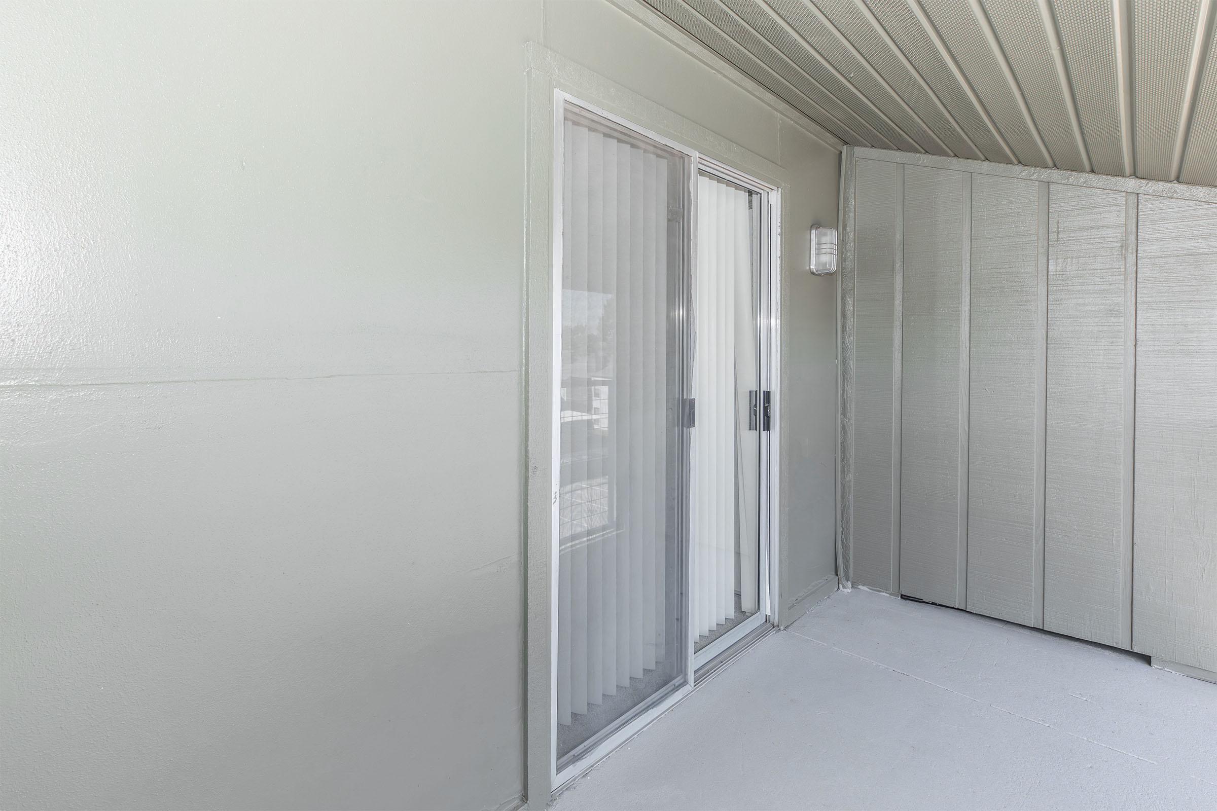 A view of a light gray exterior wall with sliding glass doors. The doors are partially covered by sheer vertical blinds, and the surrounding wall is smooth and untextured. The floor appears to be concrete, and the area is well-lit, suggesting it is outdoors or on a balcony.