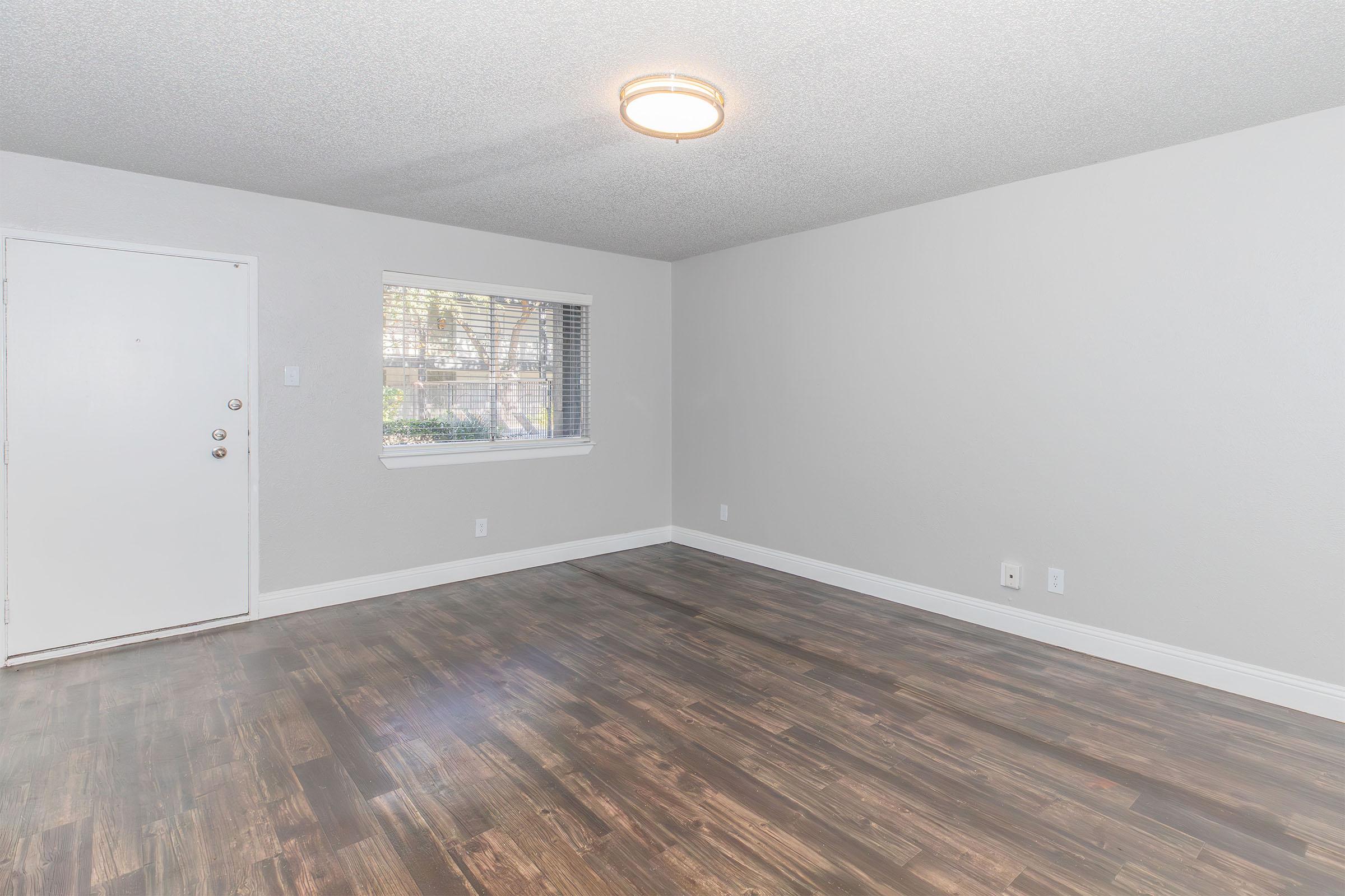 Empty room with freshly painted light gray walls and wooden flooring. A window with blinds allows natural light to enter, and a simple ceiling light fixture illuminates the space. The room has a plain white door on one side and is devoid of furniture, creating a clean, minimalistic atmosphere.