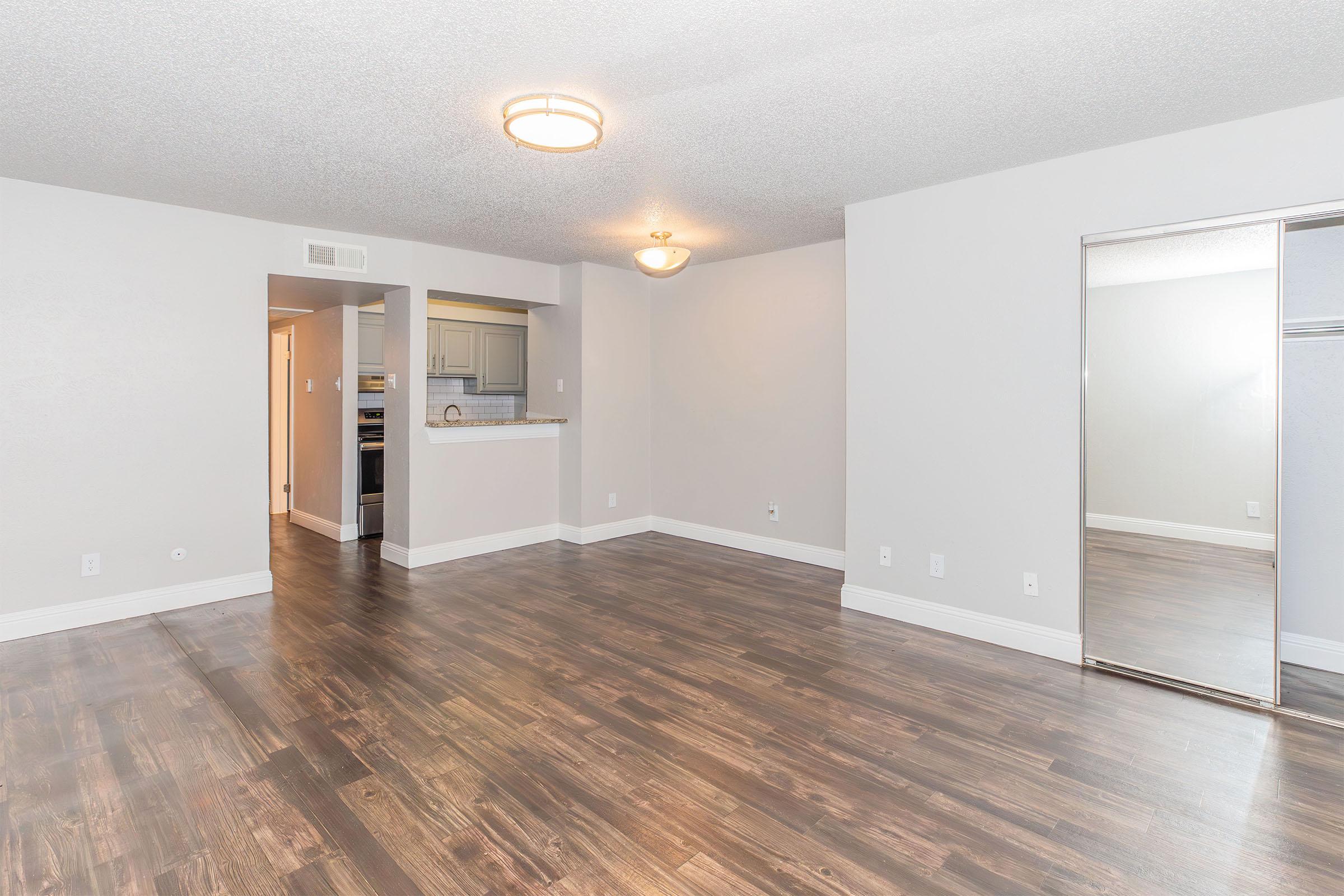 A spacious, empty living area with light-colored walls and brown wooden flooring. A doorway leads to a kitchen area in the background, and a large mirror is mounted on one wall. The room is well-lit with overhead lighting and has a clean, minimalist aesthetic.