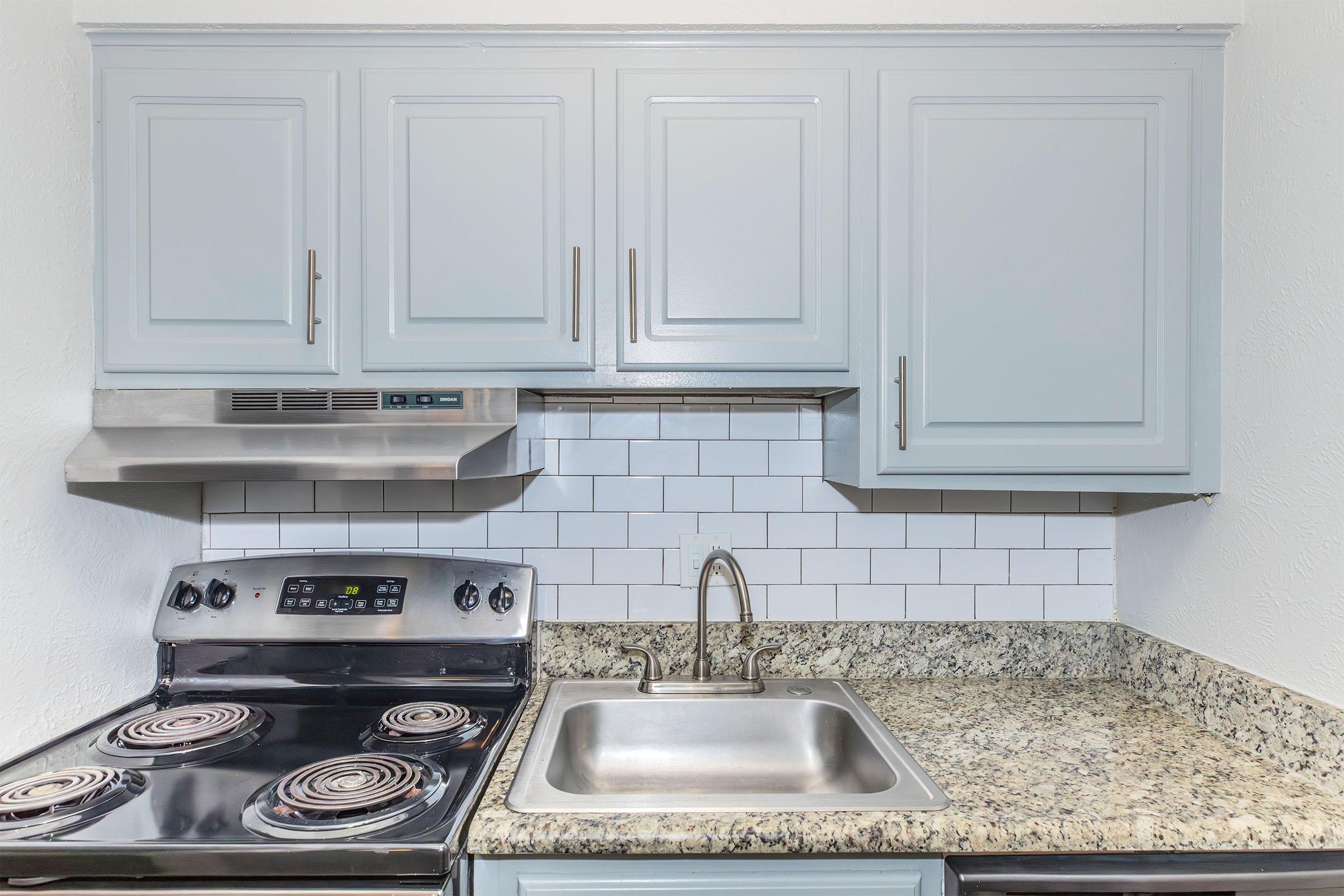 A modern kitchen with light blue cabinets, stainless steel appliances, and a gray granite countertop. The stove has four burners, and there is a stainless steel sink with a faucet. White subway tiles are used as a backsplash, creating a clean and contemporary look.