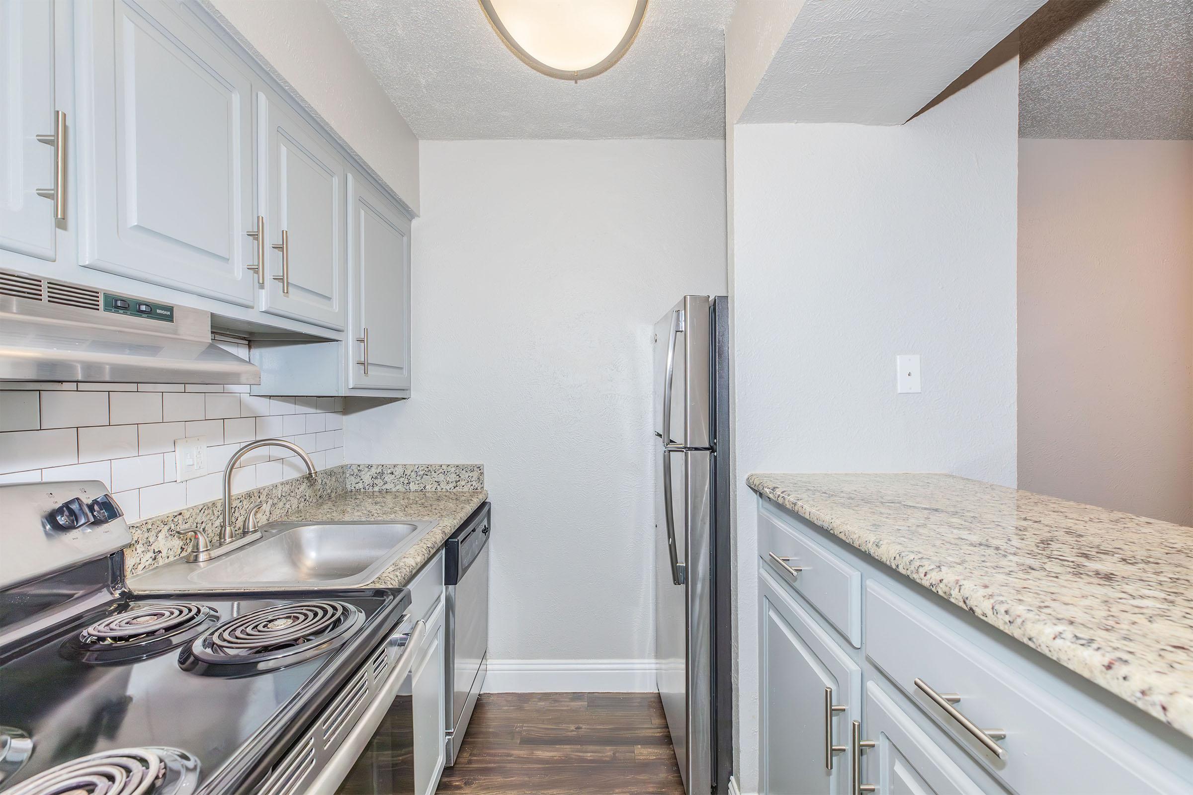A modern kitchen featuring light gray cabinets, a granite countertop, a stainless steel refrigerator, and a black stove with an overhead microwave. The space has a neutral-colored wall, and the flooring appears dark wood, creating a cozy yet contemporary atmosphere.