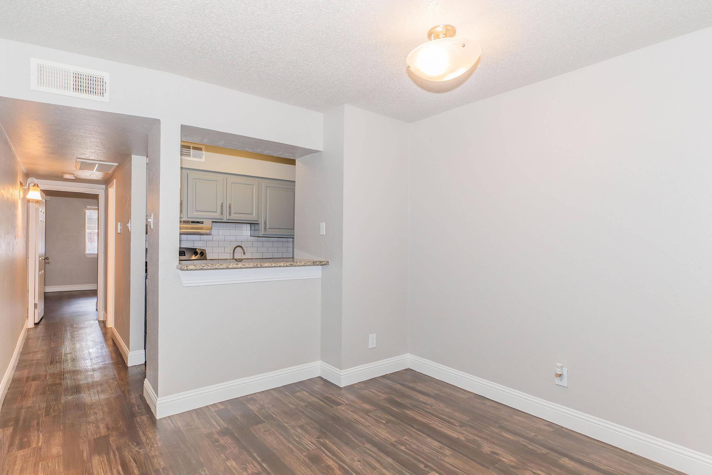 Bright and empty interior of an apartment with light-colored walls and dark wood laminate flooring. A small kitchen area is visible in the background, featuring light cabinets and a countertop. A hallway can be seen leading to another room, illuminated by a ceiling light.