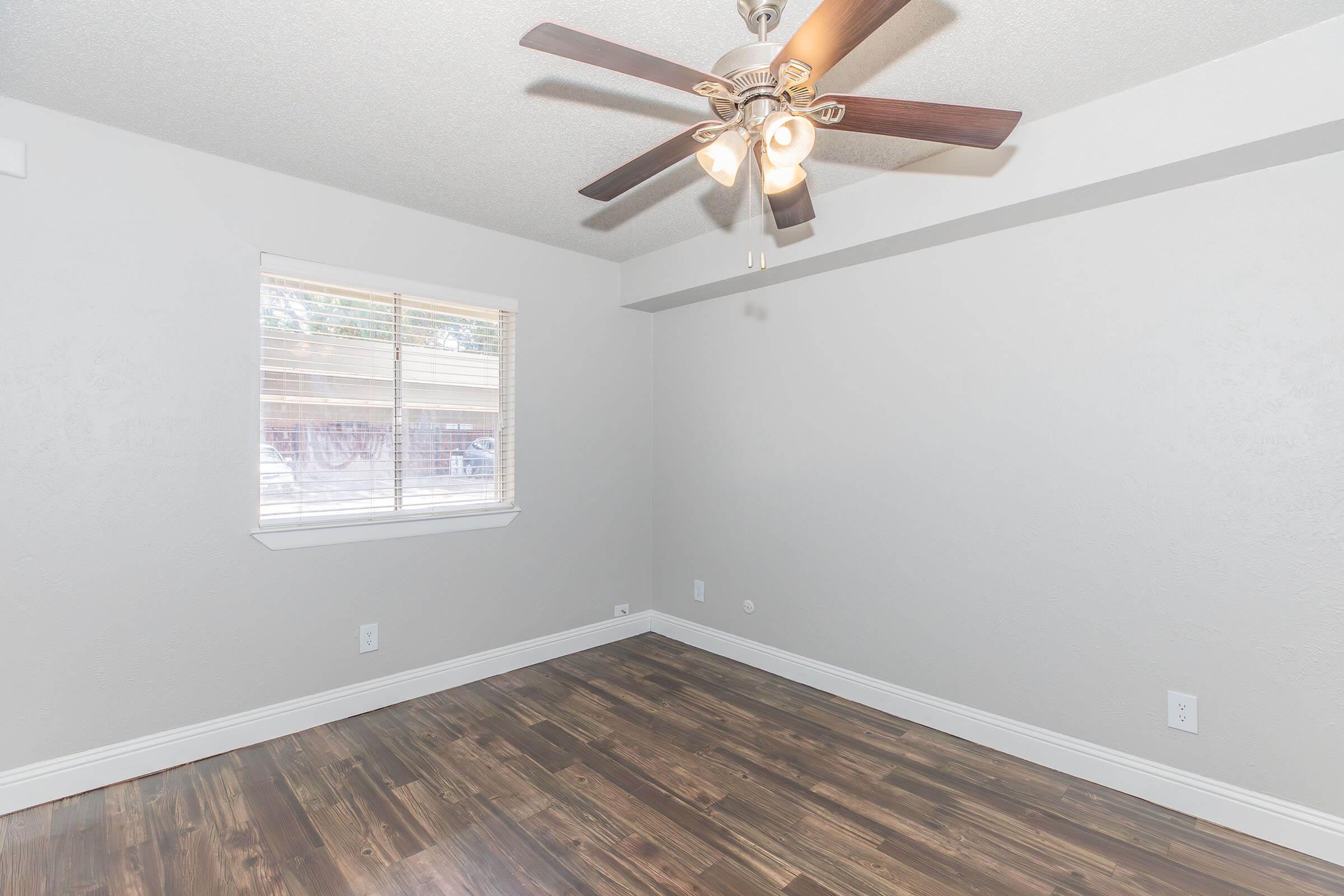 A bright, empty room with light gray walls and dark wooden flooring. A ceiling fan with four lights hangs from the ceiling, and there is a window allowing natural light to enter. The room appears spacious and clean, suitable for various purposes.
