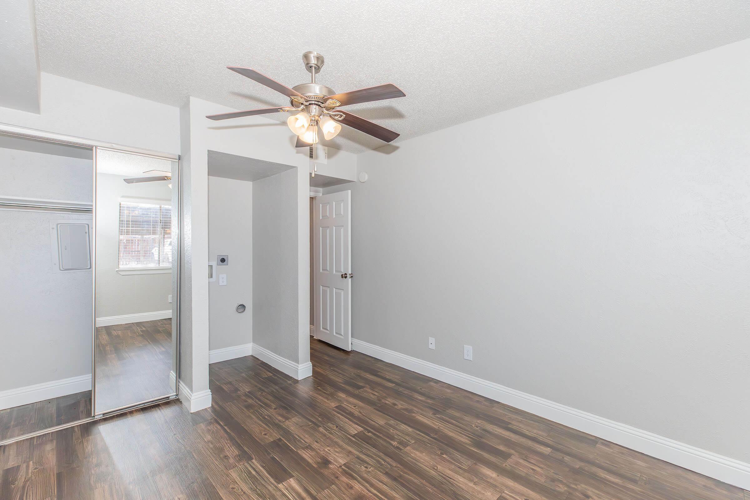A spacious and well-lit room featuring a wooden floor, a ceiling fan with lights, and a mirrored closet door. The walls are painted in a light color, and there is a door leading to another area. Natural light enters through a window, enhancing the room's brightness.