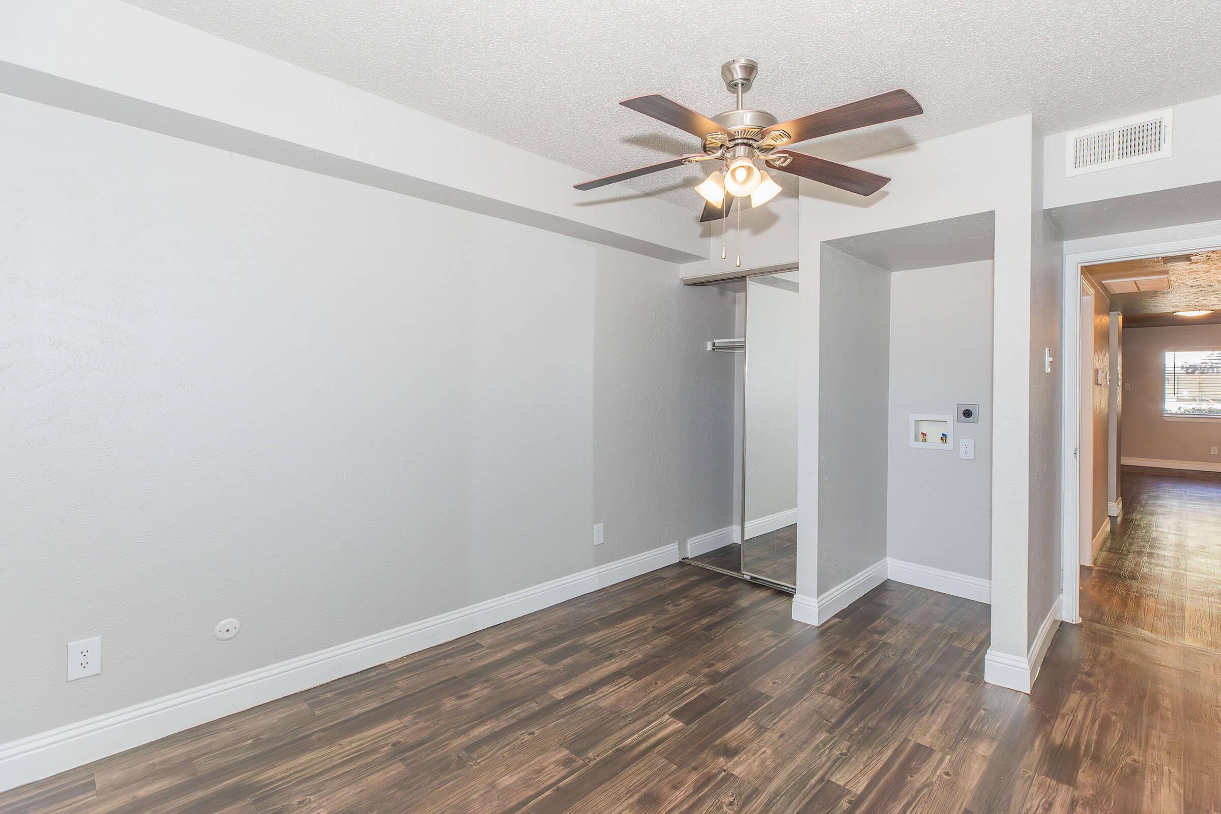 Interior of a modern living space featuring a ceiling fan, light gray walls, and hardwood flooring. There is a mirror closet on the left side and a hallway leading to another room on the right. The overall ambiance is bright and contemporary.