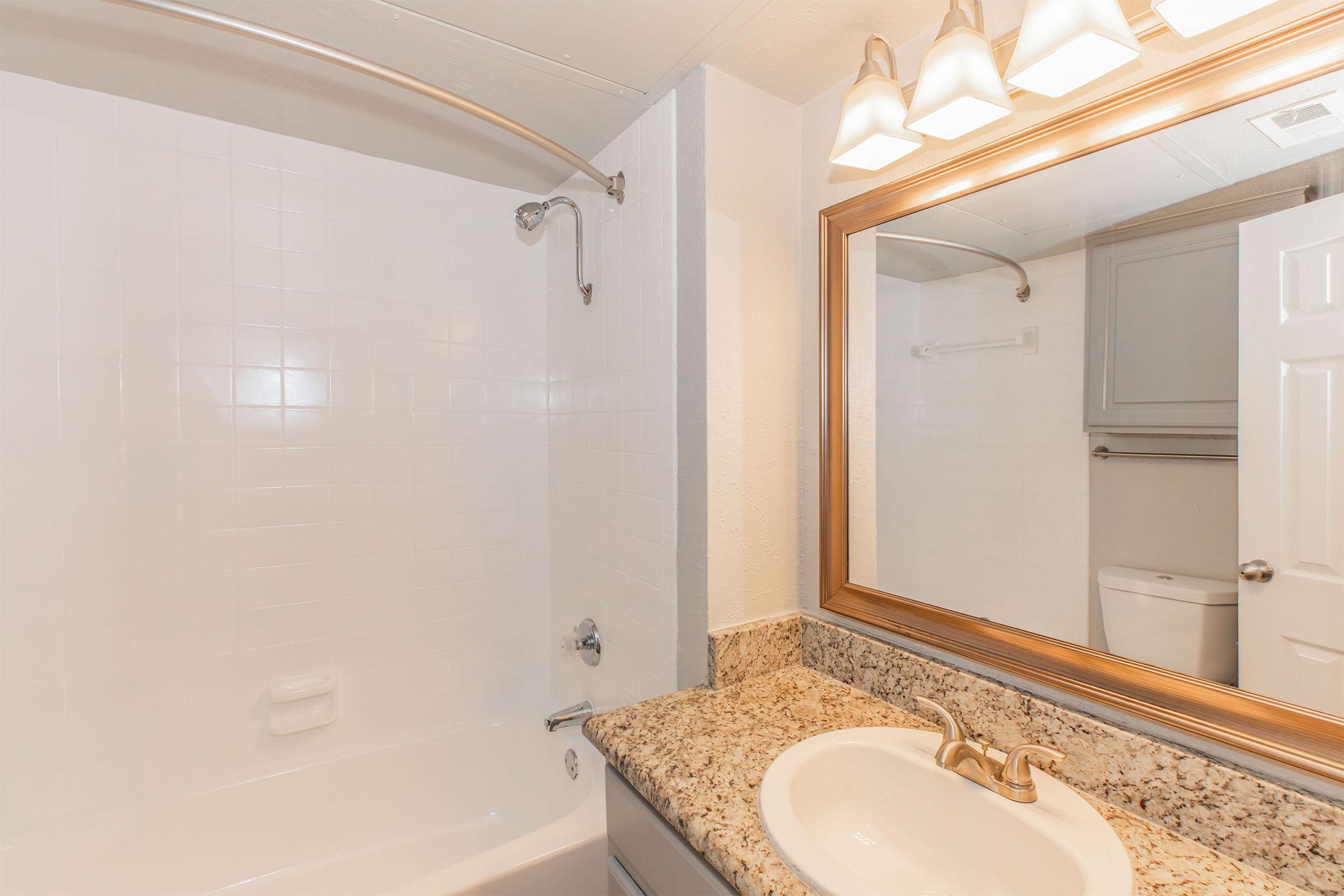 A clean bathroom featuring a white tiled shower and bathtub, a granite countertop with a sink, and a mirror above. The lighting is provided by three fixtures above the mirror. A white toilet is visible in the background, and there are towel racks and a light-colored cabinet.
