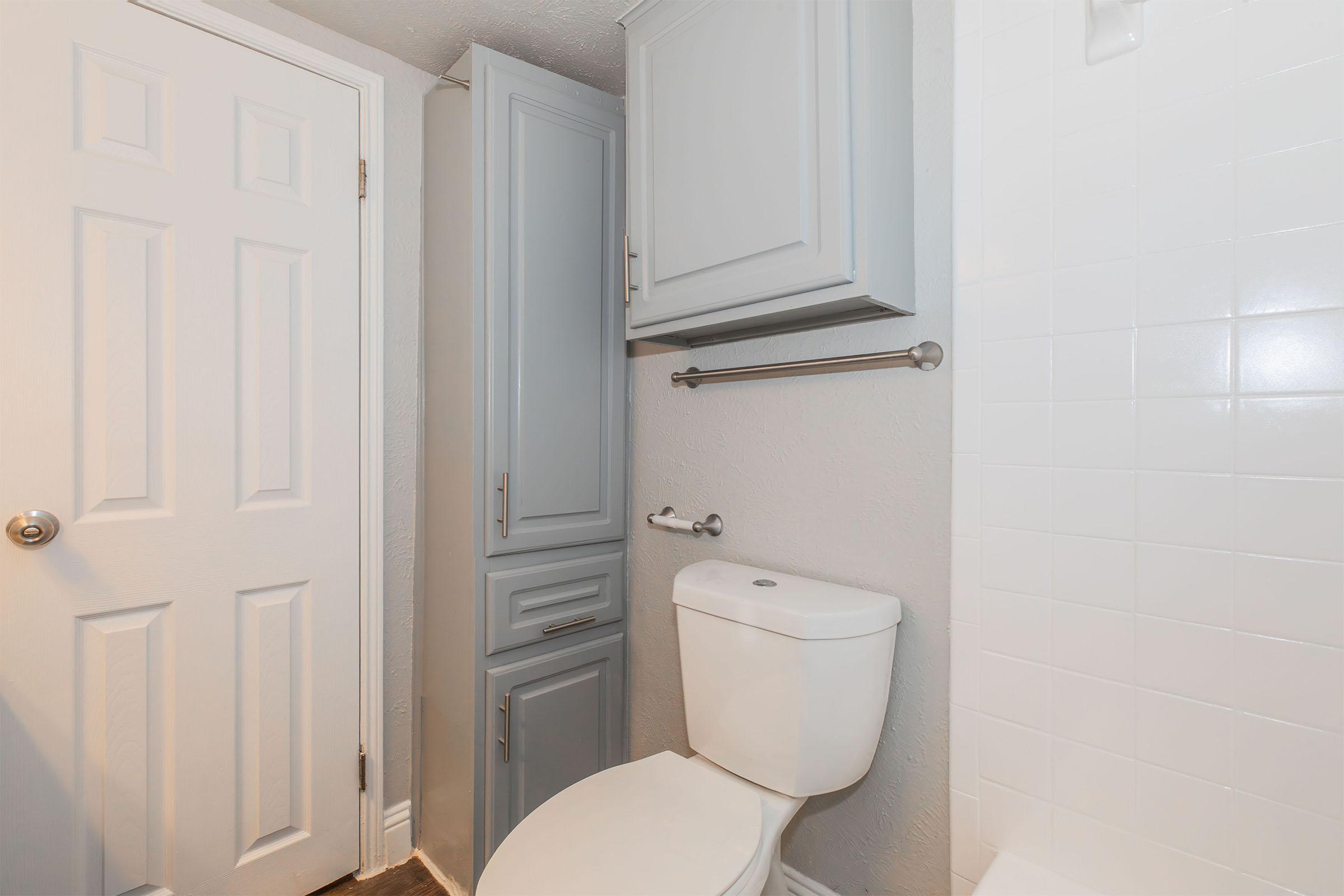 A small bathroom featuring a white toilet, light gray cabinets, a towel bar, and a white tiled shower. The door is closed, with a neutral-colored wall providing a clean and simple aesthetic.