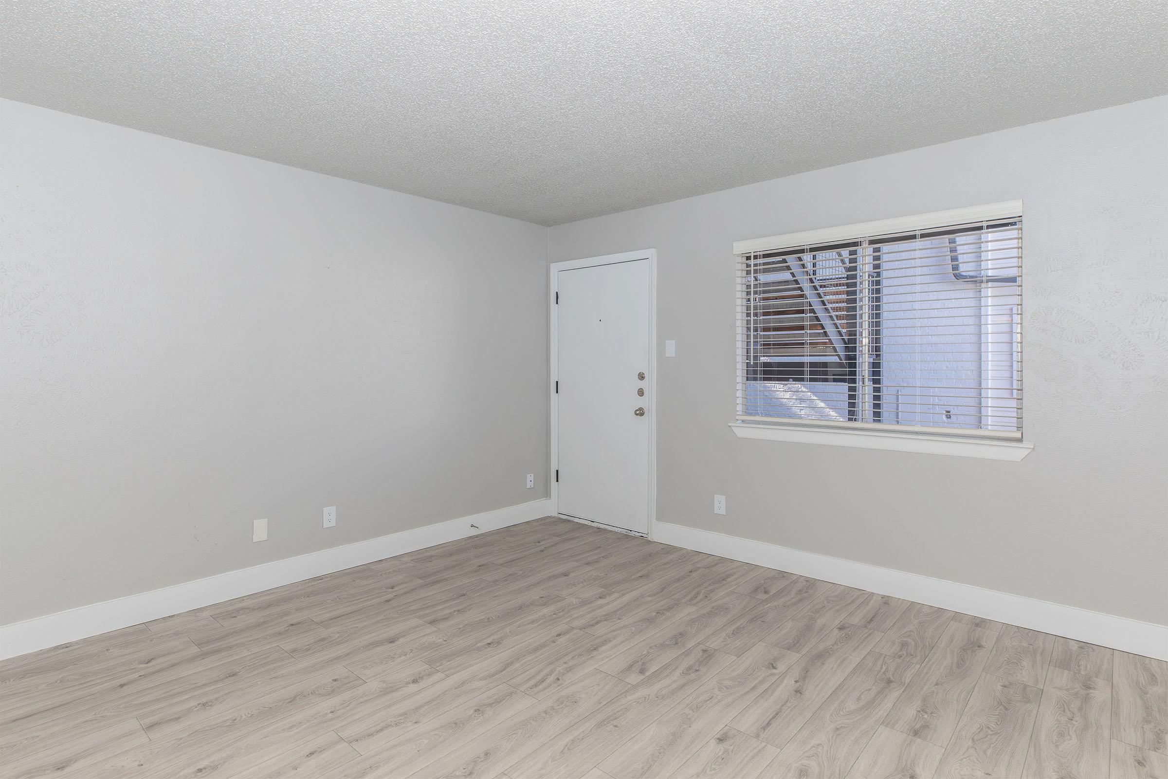 Empty room with light gray walls, a door on the left, and a window on the right featuring blinds. The flooring is light-colored wood laminate, and the overall space appears tidy and well-lit. No furniture or decorations present.