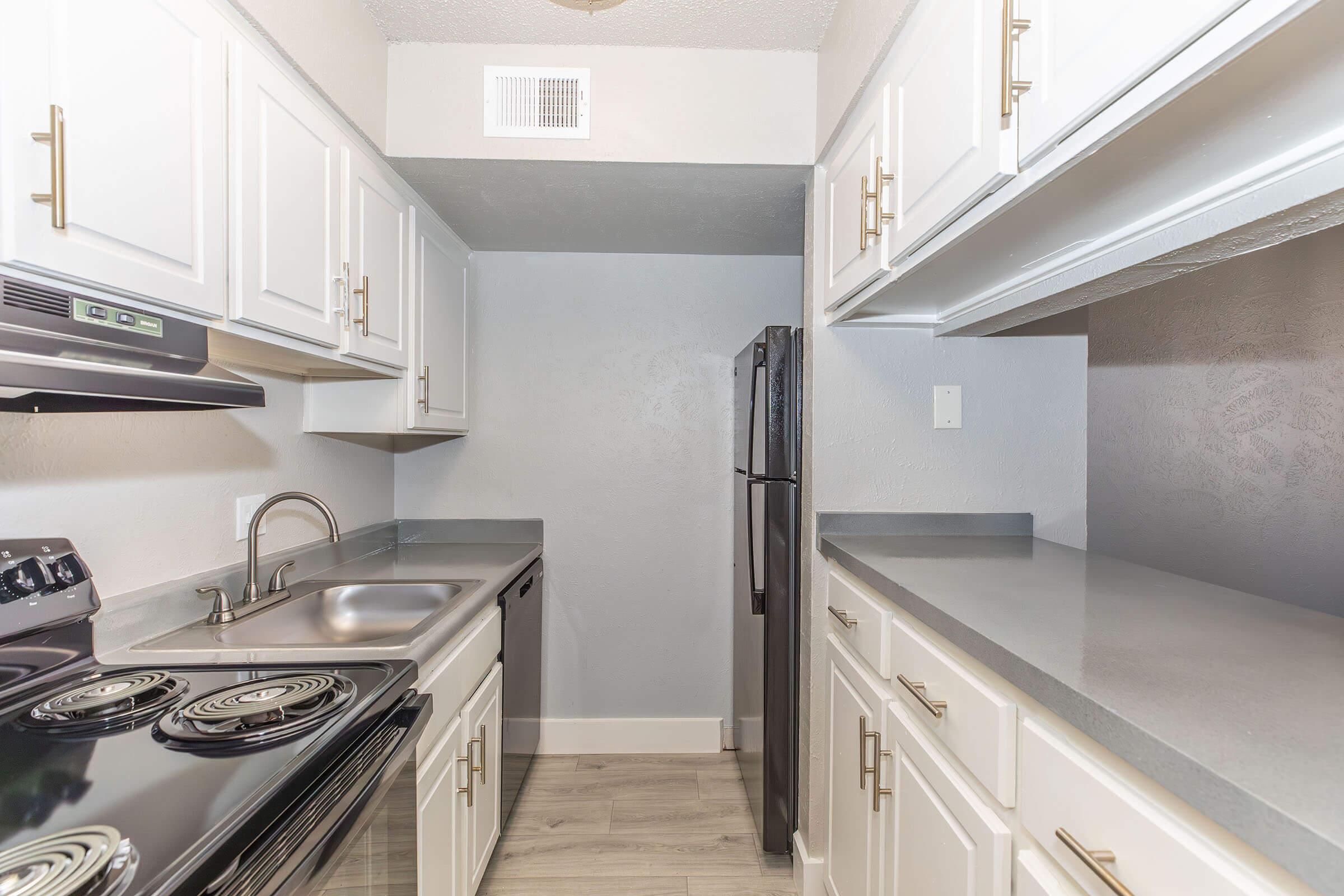 Modern kitchen featuring white cabinetry with gold hardware, a stainless steel sink, and black appliances including an electric stove and refrigerator. Gray walls and laminate flooring create a bright, contemporary atmosphere. Ample counter space and open shelving enhance functionality.
