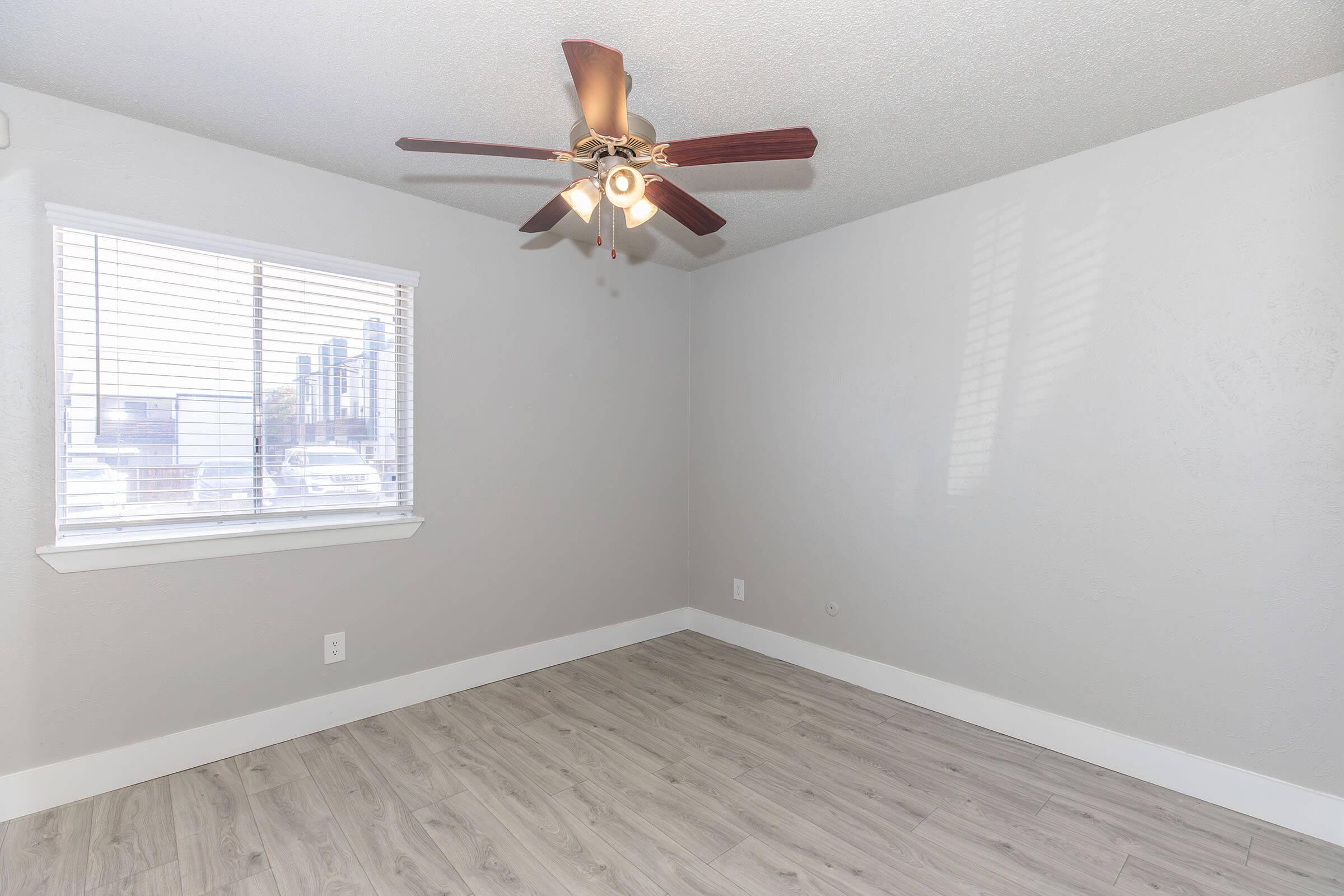 A sparsely furnished room with light gray walls, a ceiling fan with wooden blades, and a window with blinds. The flooring is light-colored wood-like material, and there is minimal furniture or decoration, emphasizing a clean and open space.