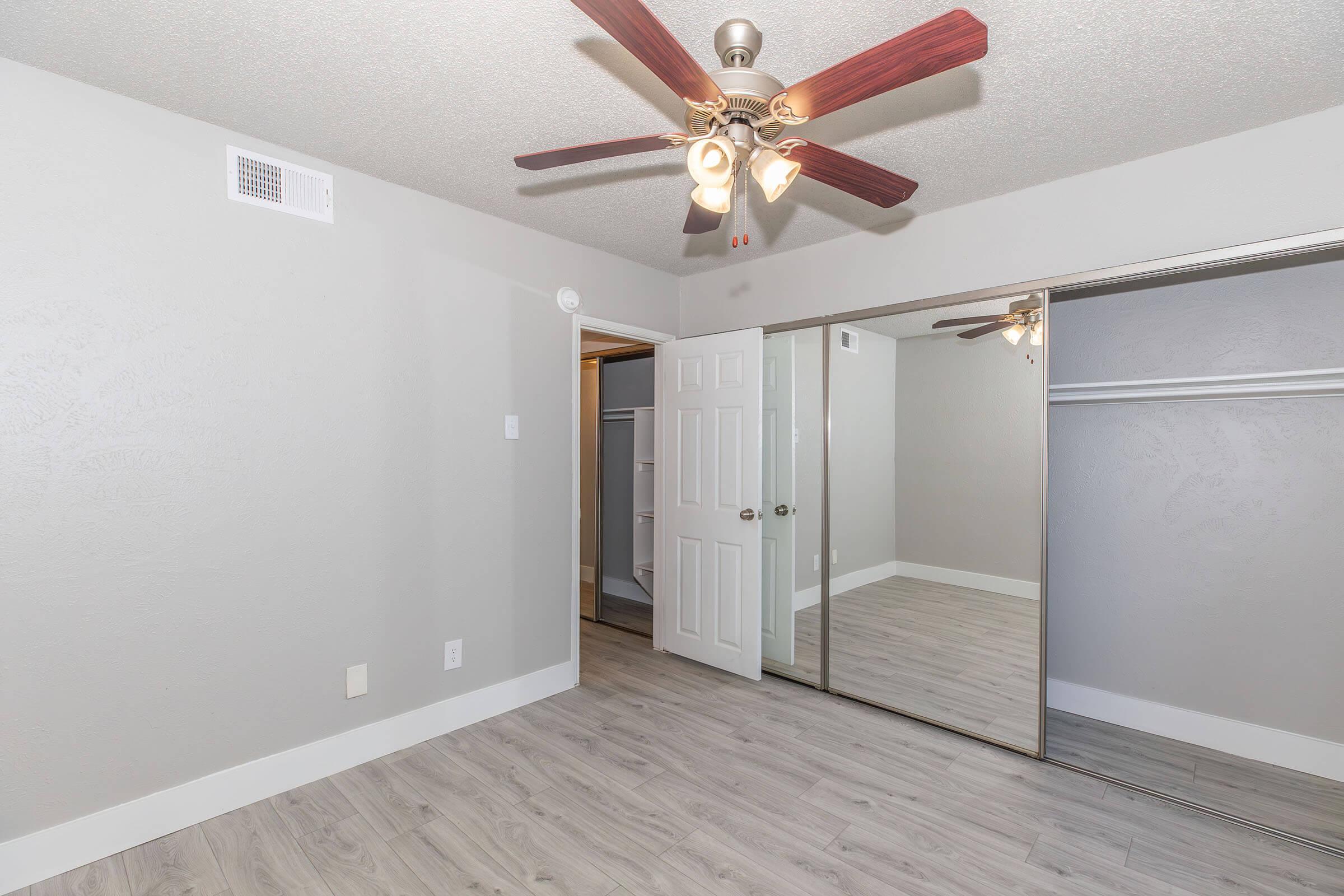 A well-lit bedroom featuring a ceiling fan with brown blades, light gray walls, and a mirrored closet. The flooring is a light wood laminate, and there is a doorway leading to another room. Overall, the space appears modern and freshly updated.