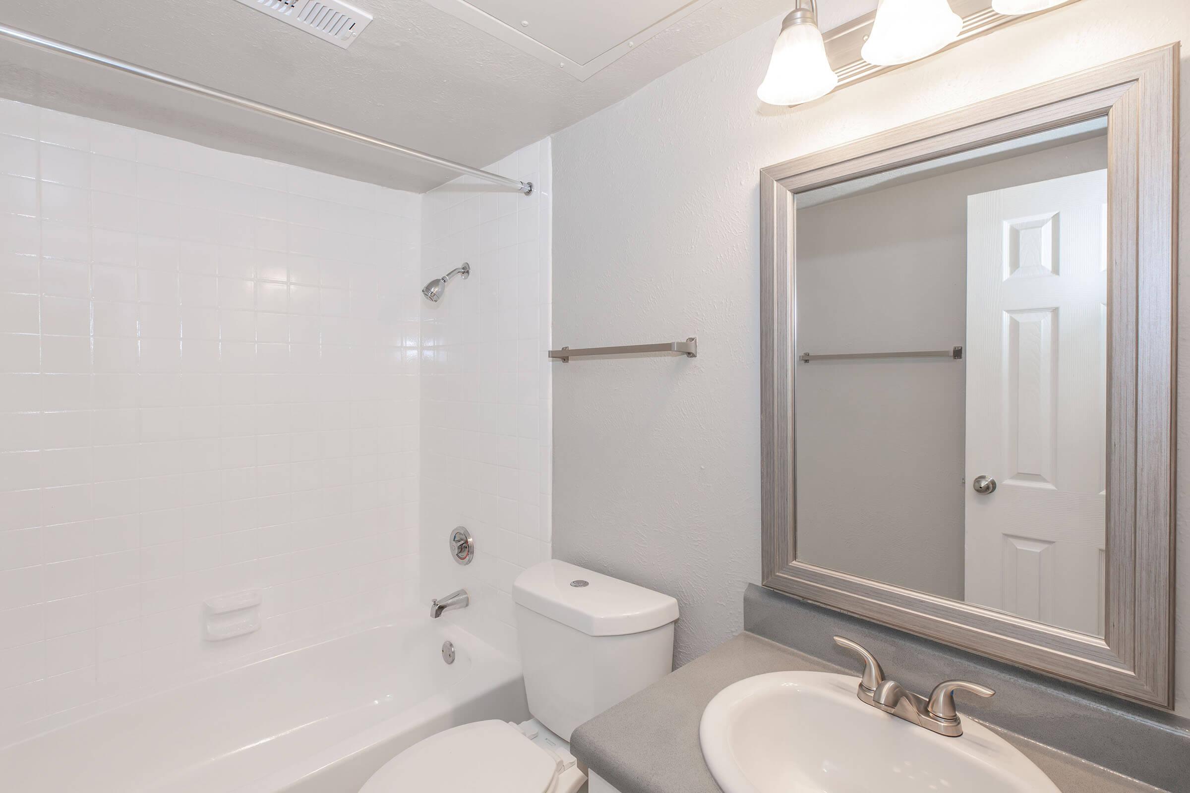 A clean, modern bathroom featuring a white bathtub and shower combination, a toilet, and a sink with a countertop. A large mirror with a wooden frame hangs above the sink, while a door is visible in the background. The walls are painted light gray, and there are two light fixtures above the mirror.
