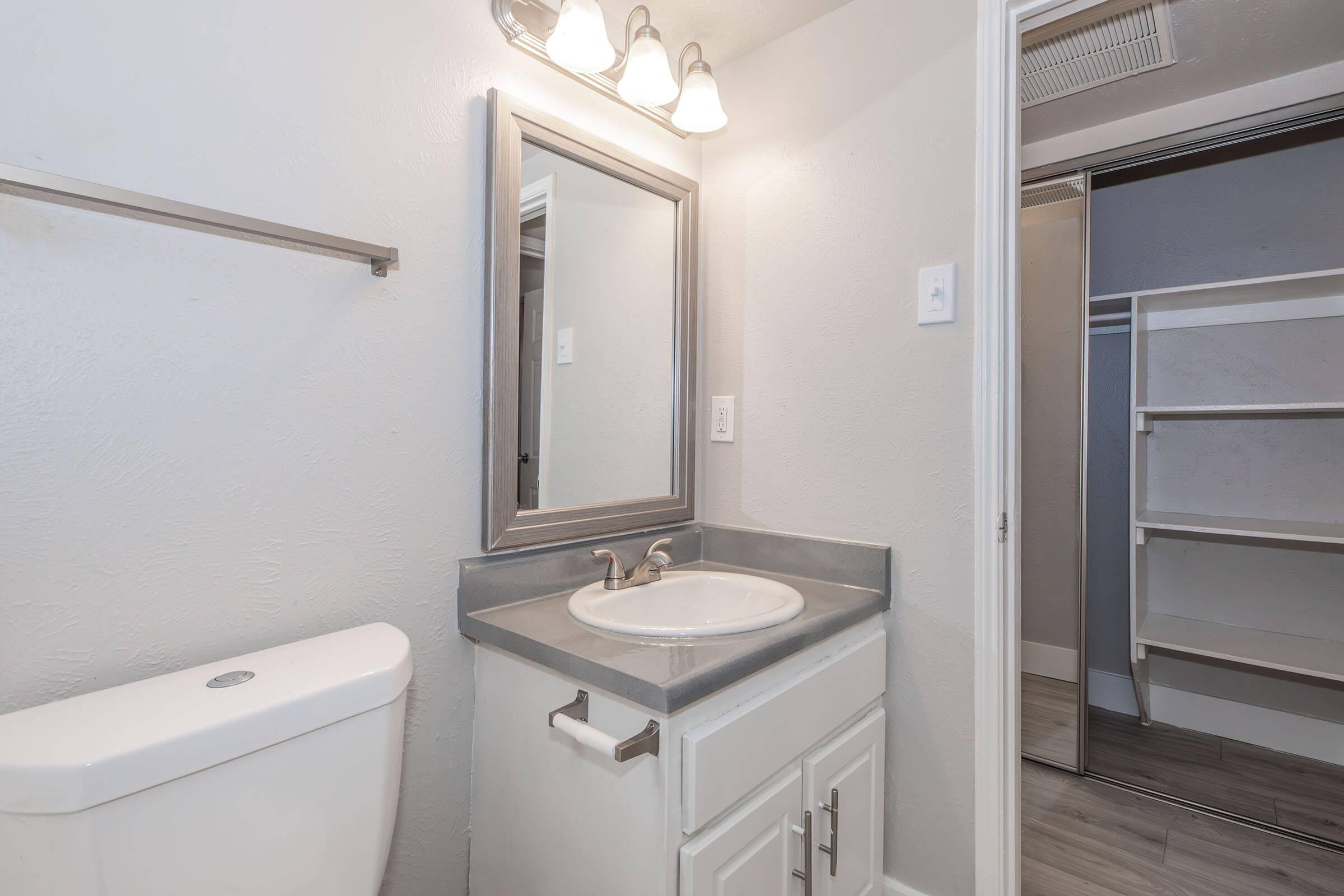 A modern bathroom featuring a white toilet, a gray countertop with a sink, a mirror above the sink, and a light fixture. The walls are light gray, and a closet with sliding doors is visible on the right, showcasing shelves inside. The overall design is clean and minimalist.