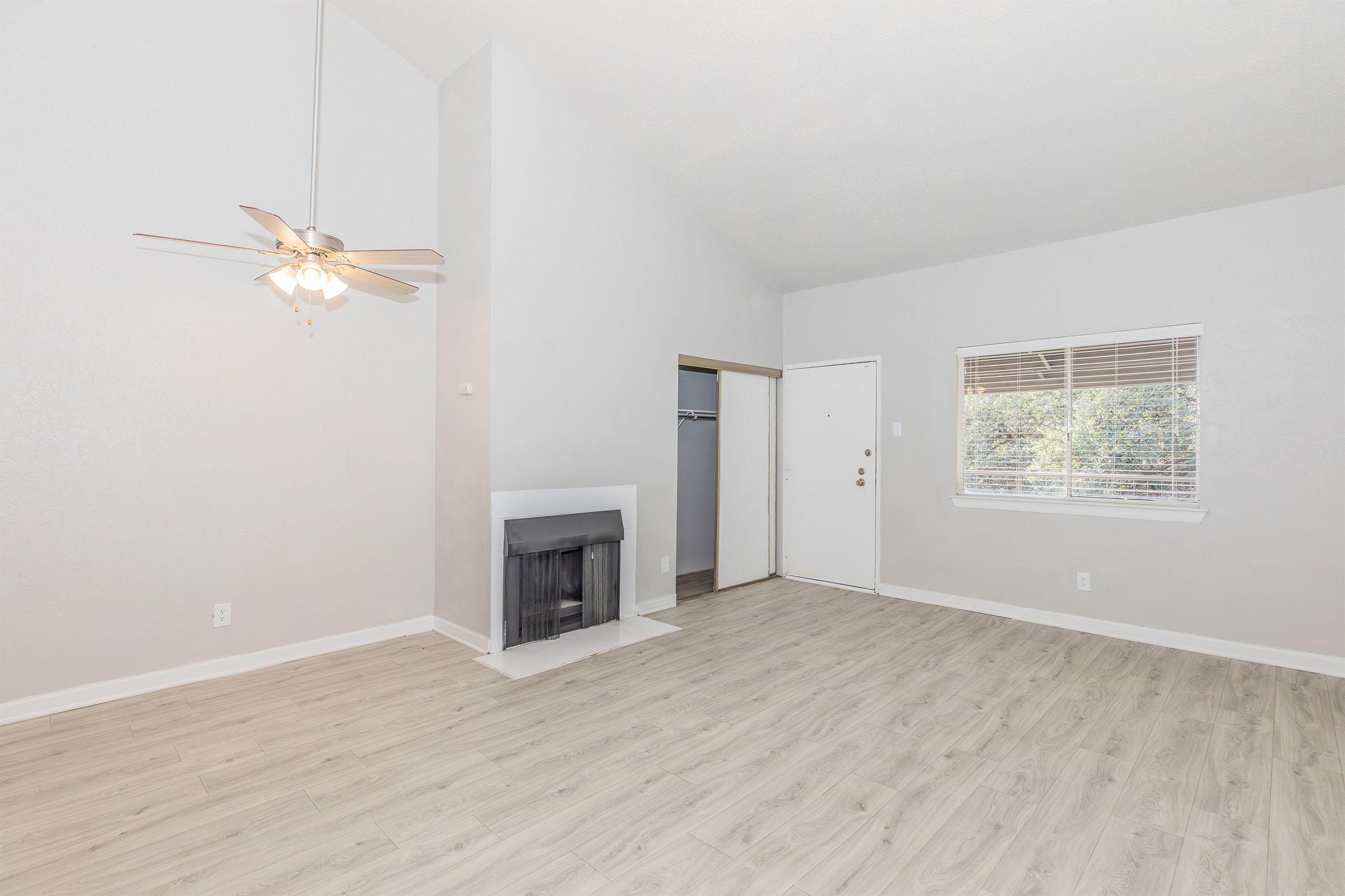 Spacious living room featuring light-colored walls, a ceiling fan, and wood-like flooring. A fireplace is present, along with a doorway leading outside. A closet can be seen in the background, and there is a large window allowing natural light to brighten the space.