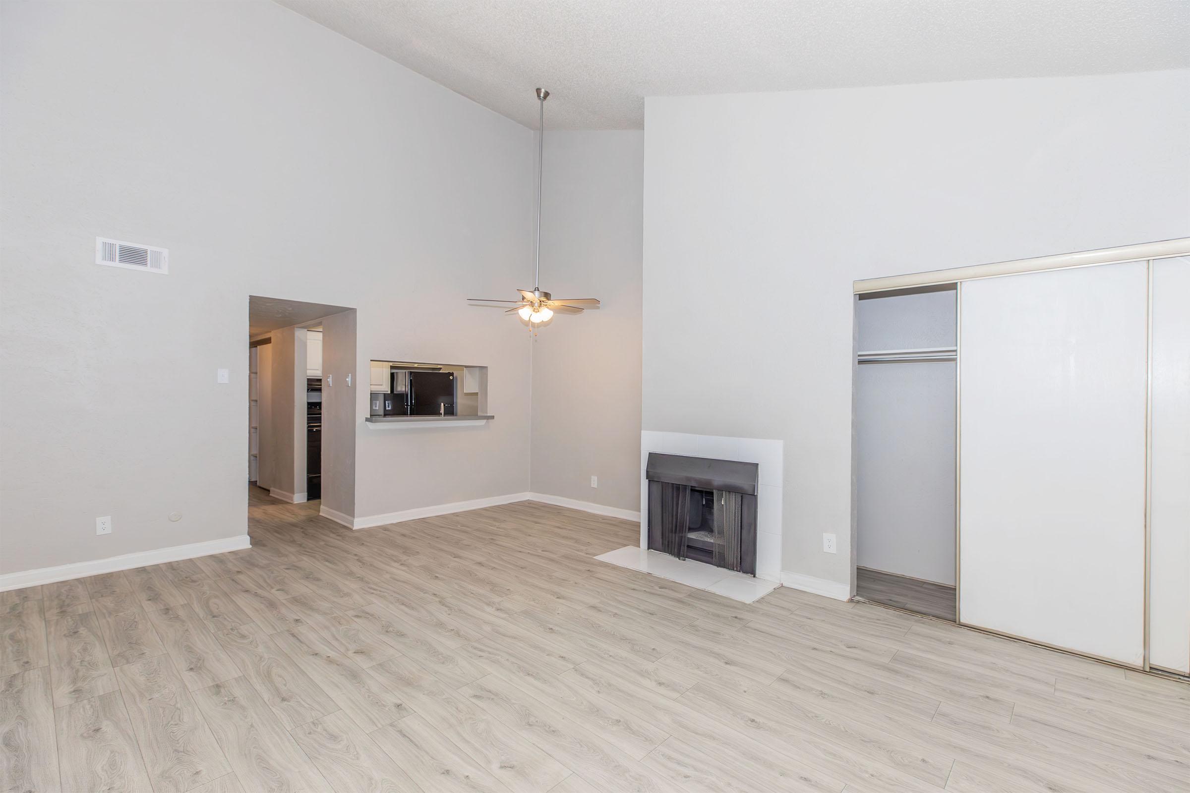 Spacious interior of a living area featuring high ceilings, light-colored walls, and wood-style flooring. Includes a ceiling fan, a fireplace, and a closet with sliding doors. A doorway leads to a kitchen area in the background, enhancing the open concept design.