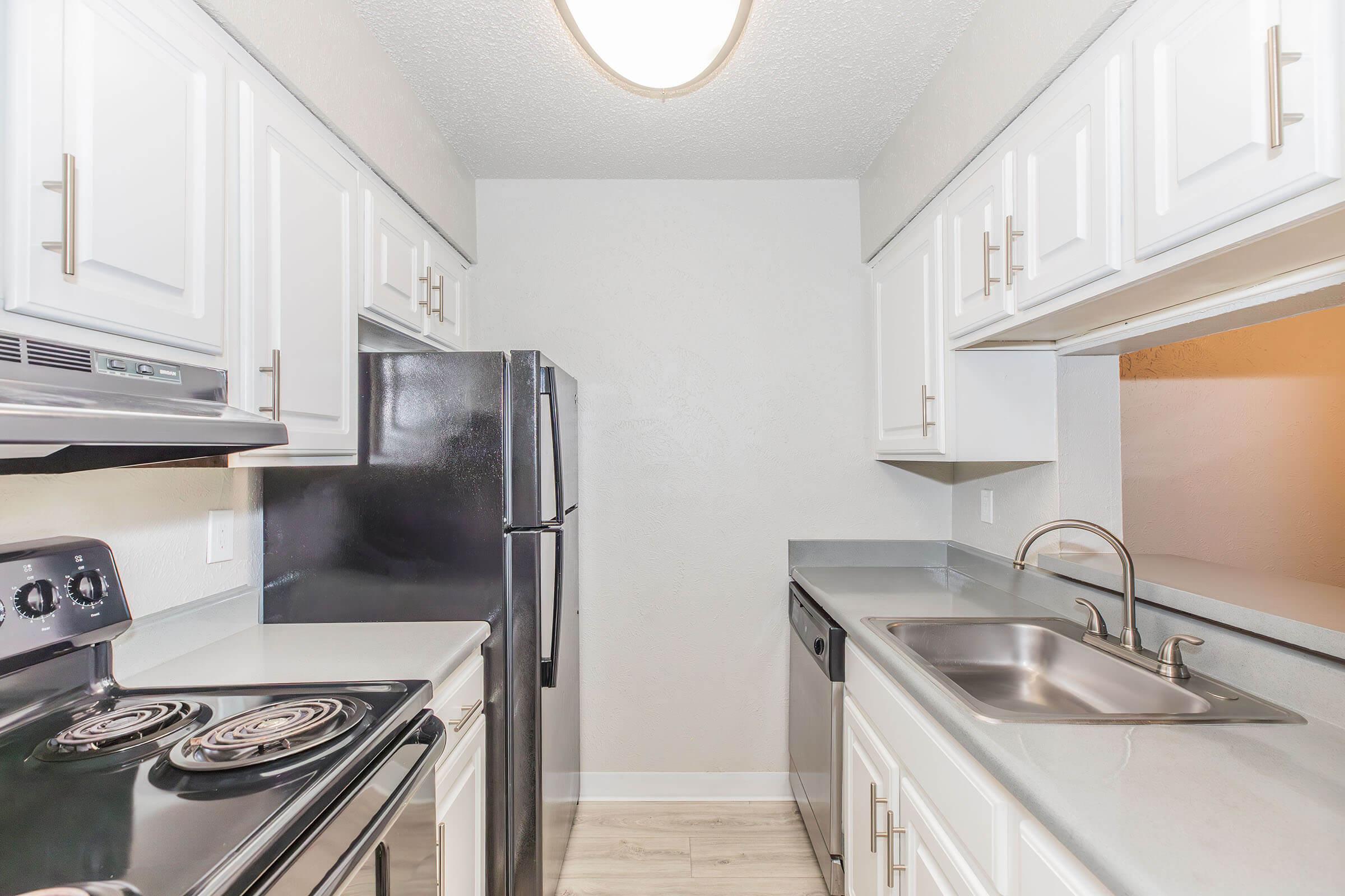 Modern kitchen featuring white cabinetry, black appliances including a refrigerator and stove, a stainless-steel sink, and light countertops. The space is well-lit with ceiling lighting, and the layout is functional with ample counter space and a dishwasher.