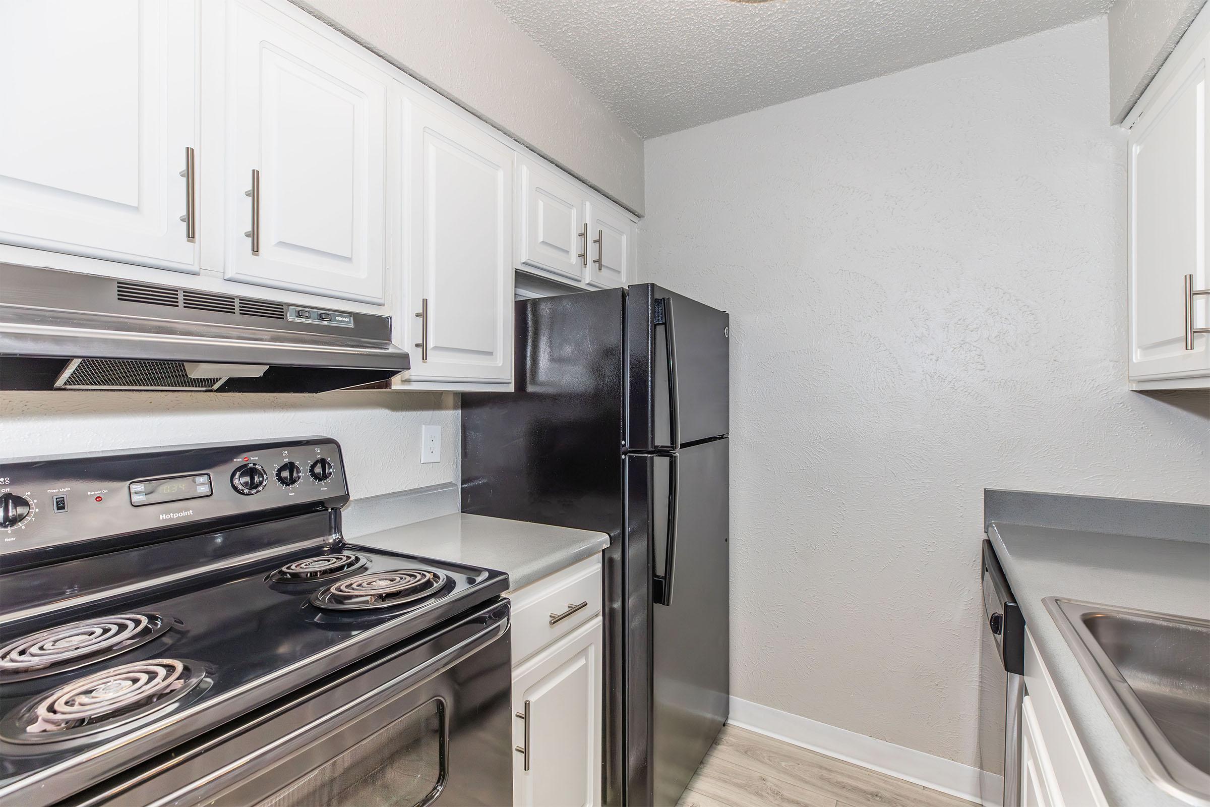Modern kitchen with white cabinets, a black refrigerator, and an electric stove with four burners. Countertops are gray, and the walls are painted in a light neutral shade. The space is well-lit and features a clean, contemporary design.