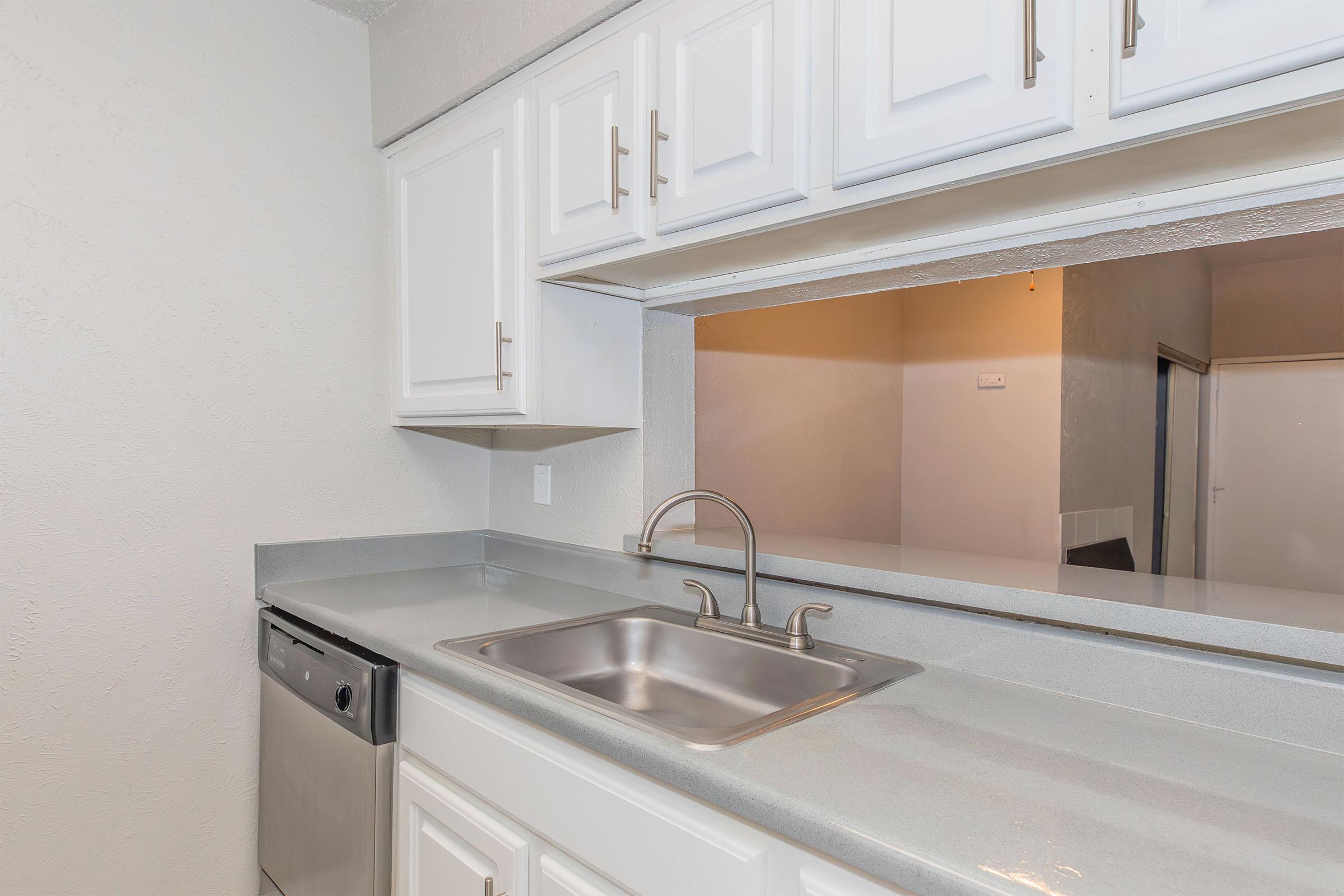 Modern kitchen with white cabinetry, a stainless steel sink, and a silver faucet. The countertop is gray and appears to be made of stone. A dishwasher is visible to the left. The space has a light-colored wall and an open view into another room, providing a bright and airy feel.