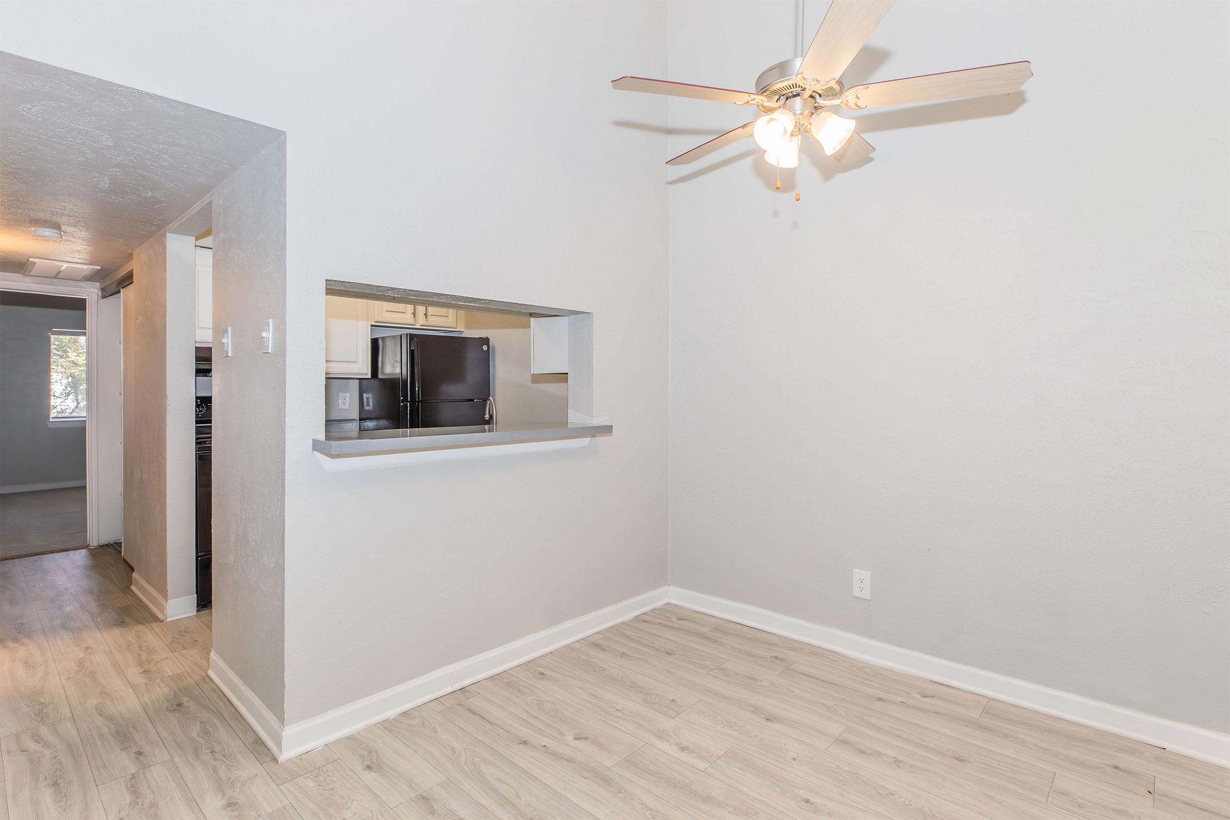 A spacious interior view of an apartment featuring light-colored walls, a ceiling fan with four bulbs, and a wooden floor. In the background, a kitchen area is partially visible through an open counter, showcasing modern appliances. Natural light enters from a nearby window, enhancing the inviting atmosphere.
