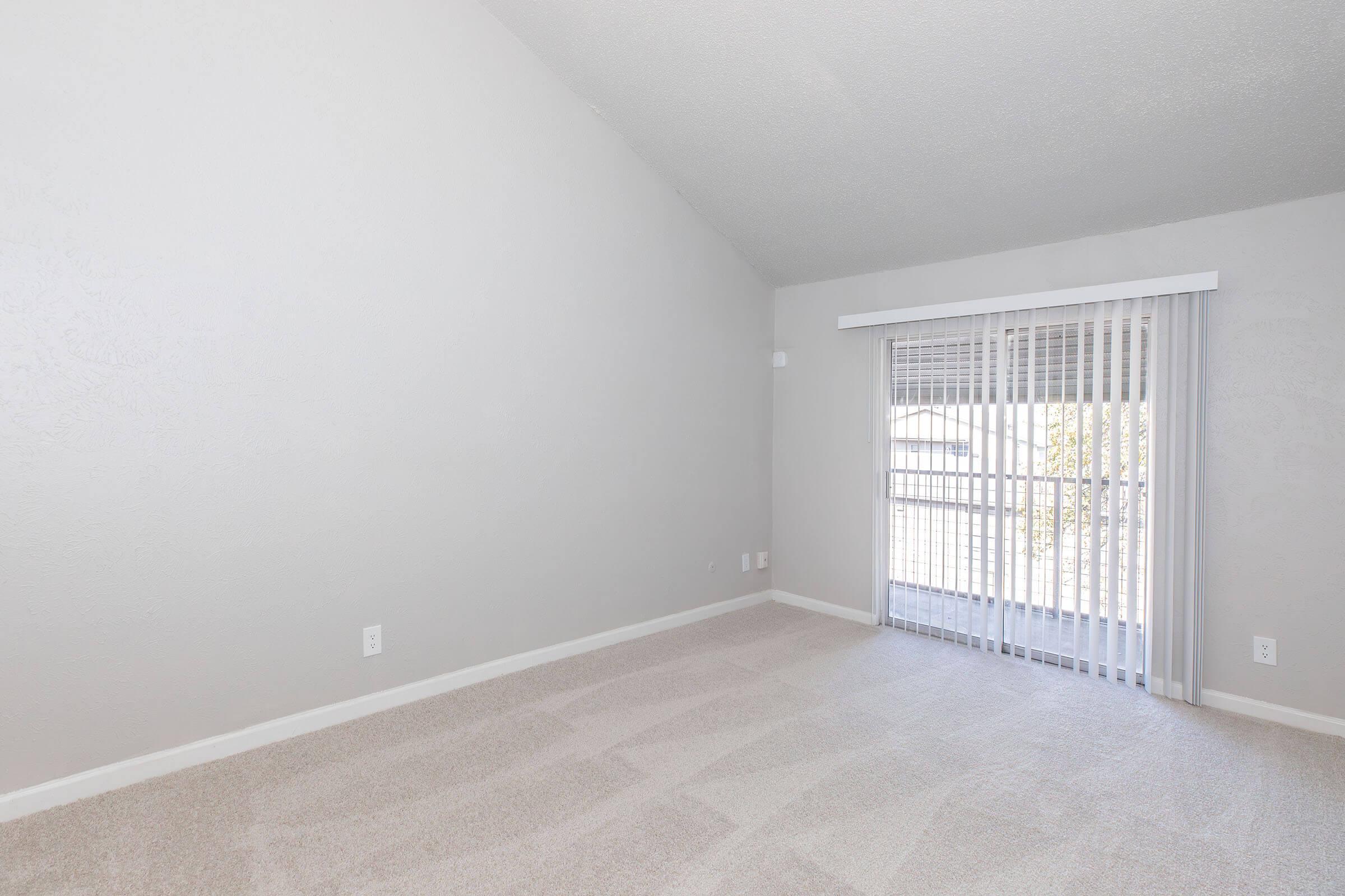 Empty room featuring light gray walls and carpet, with a tall ceiling. Large window with vertical blinds allows natural light, and a sliding door leads to a balcony or patio area. The space is unfurnished, creating a bright and open atmosphere.