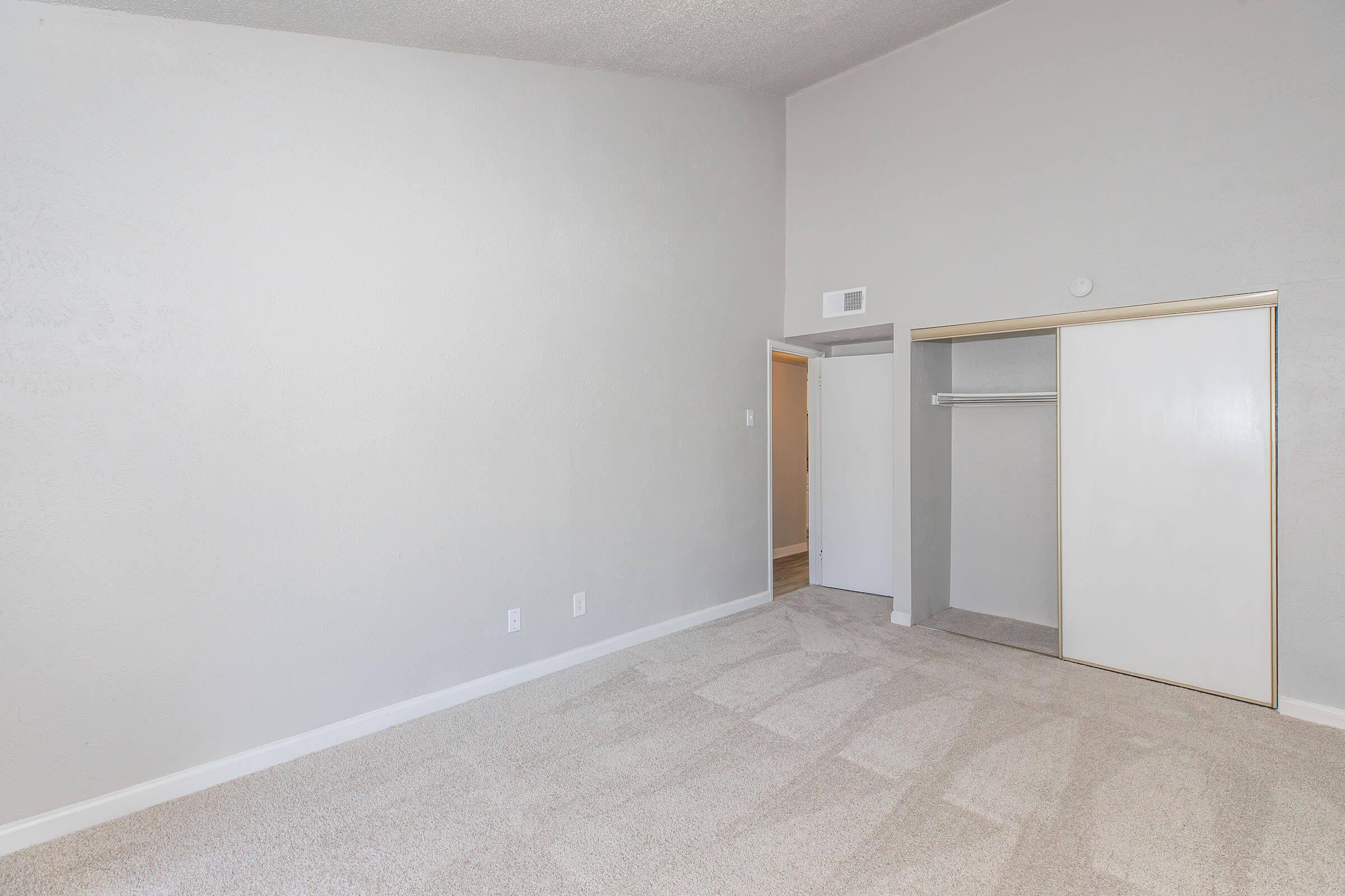 Empty room with light gray walls and plush carpet. A closet with sliding white doors is on the right, and there's an open doorway leading to another room. The ceiling is high, creating a spacious feel. Natural light fills the area, enhancing the simplicity and cleanliness of the space.