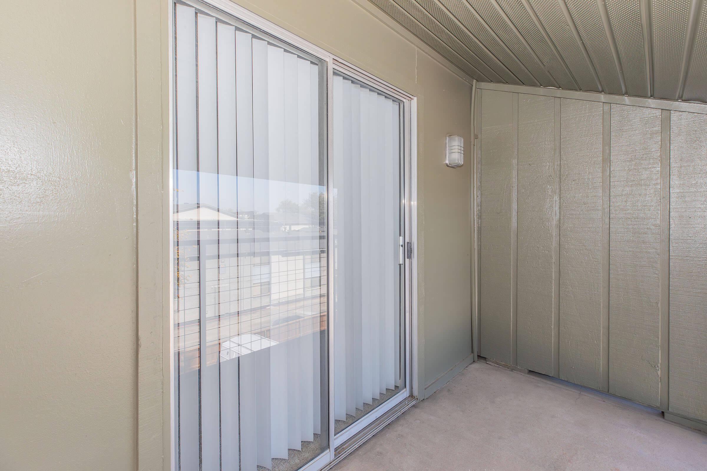 An interior view of a balcony featuring a sliding glass door with vertical blinds, surrounded by light-colored walls and a smooth floor. The space is well-lit, showcasing the simplicity and openness of the area.