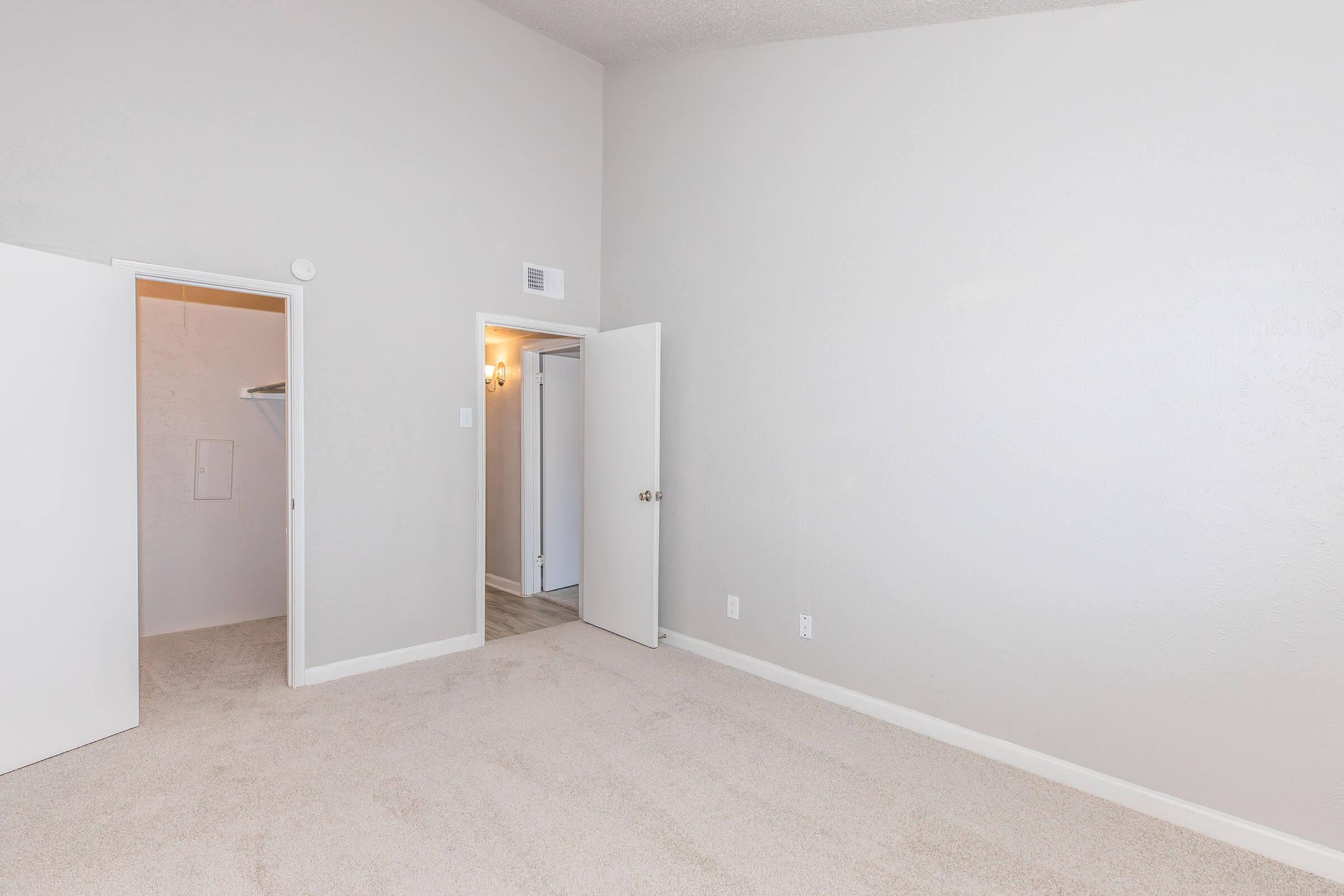 A spacious, empty room with light gray walls and soft beige carpeting. There are two open doors: one leading to a closet and the other to a hallway. Soft lighting creates a bright atmosphere, emphasizing the room's openness and potential for personalization.