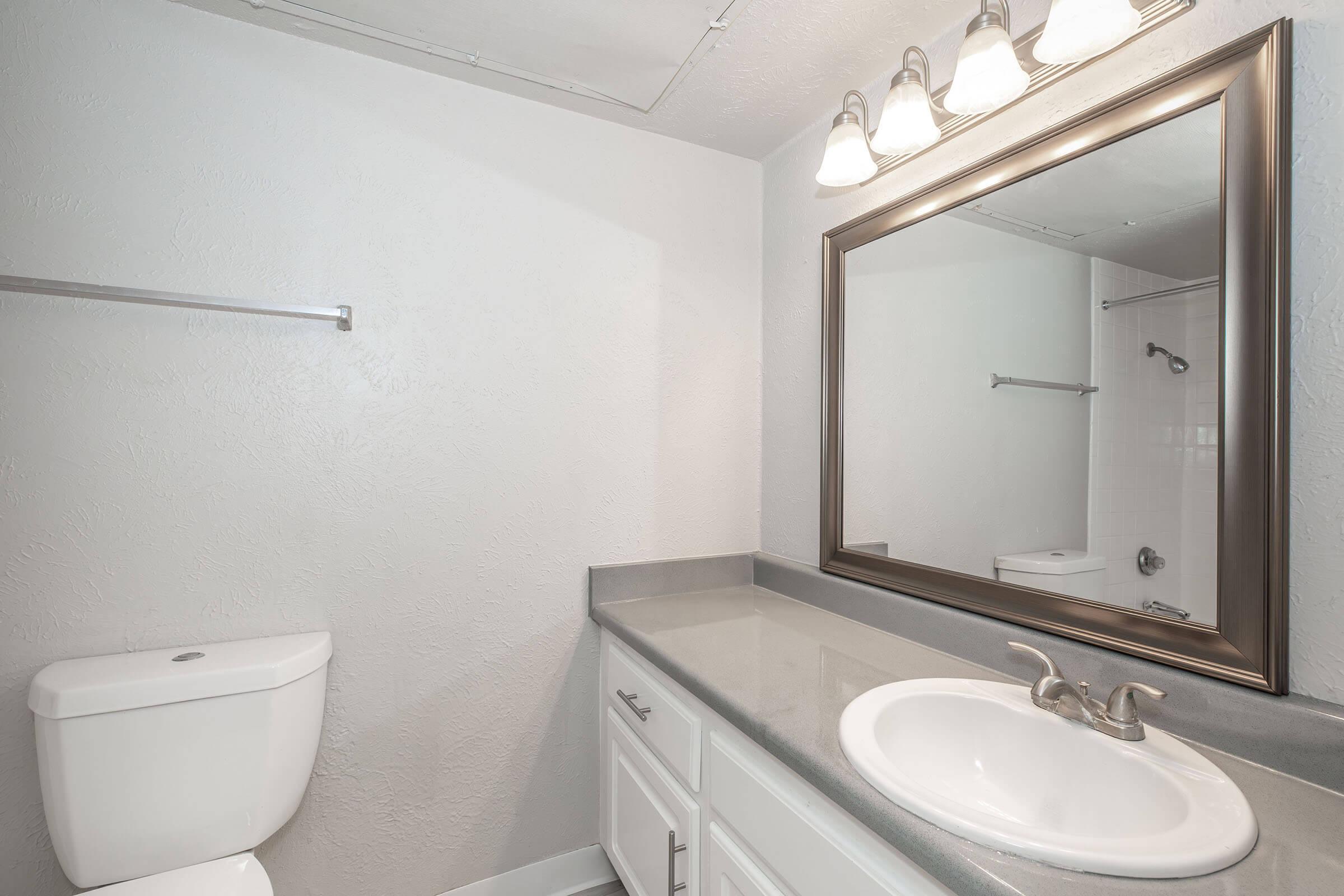 A modern bathroom featuring a white toilet, a large mirror with a decorative frame, under-sink cabinetry, a gray countertop, and three light fixtures above the mirror. The walls are painted in a light color, creating a clean and bright atmosphere. A towel rack is mounted on the wall.