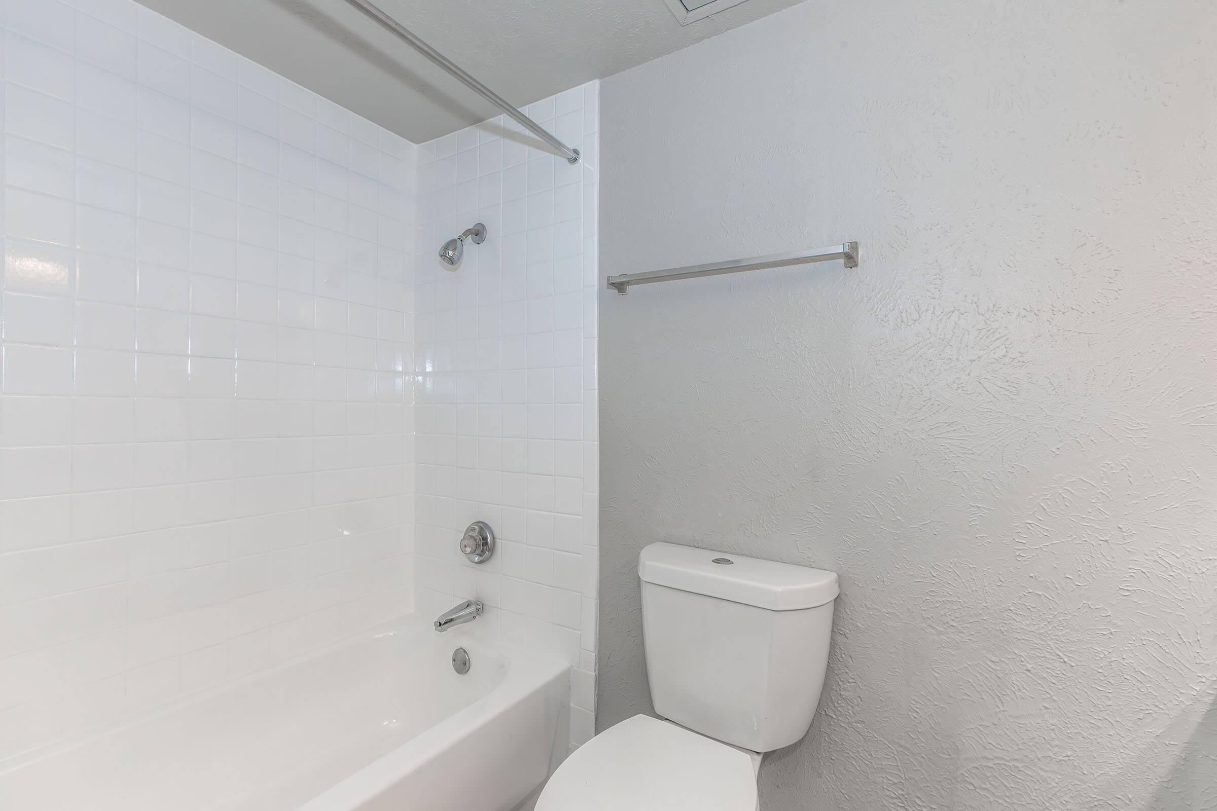 A clean, modern bathroom featuring a bathtub with a showerhead, white tiled walls, a toilet, and a towel bar. The walls are painted light gray, contributing to a bright and spacious feel.