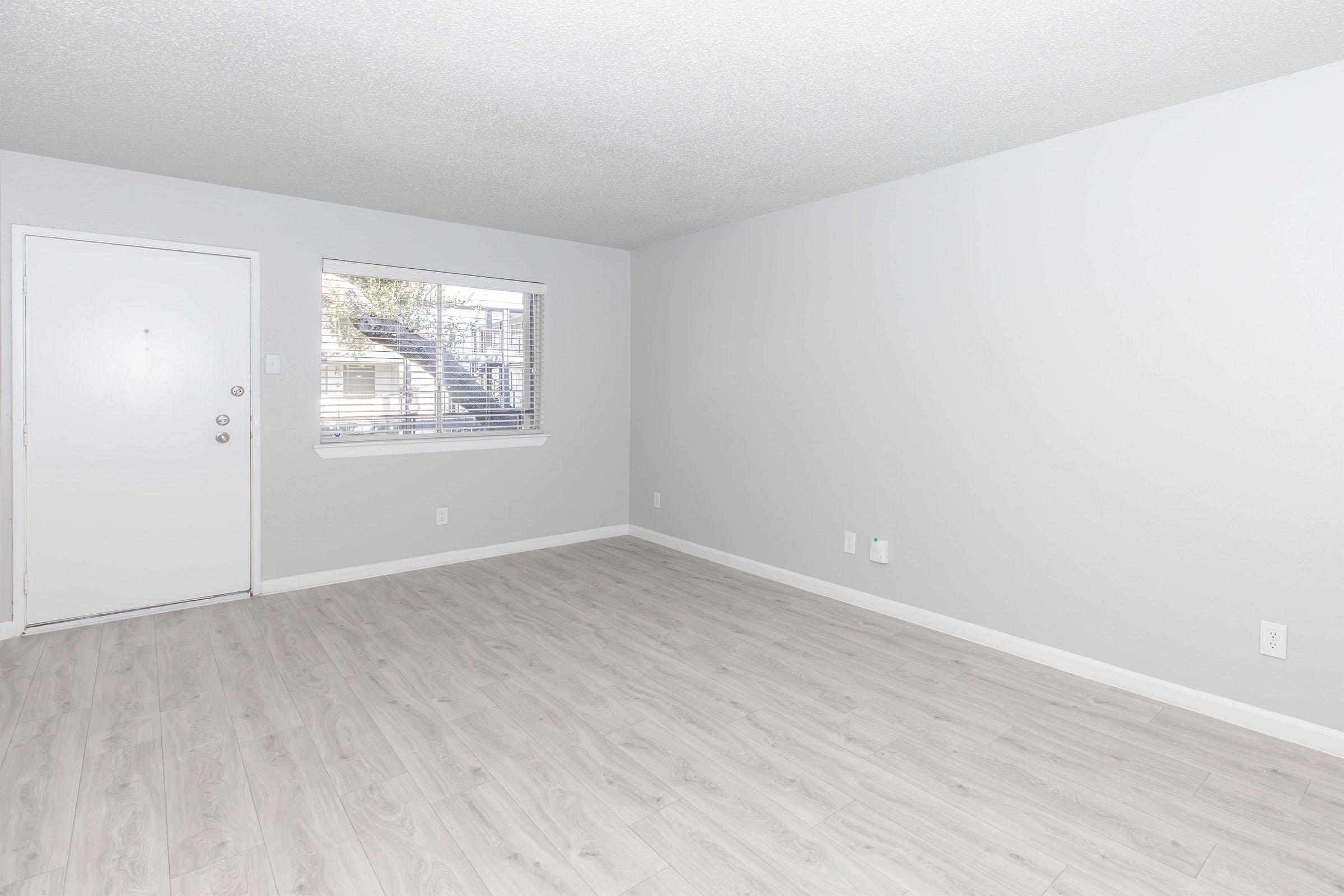 A minimalist living room with light gray walls and flooring. There's a front door on the left and a window on the right, allowing natural light to enter. The space is empty, creating a clean and airy atmosphere, suitable for various furnishings.
