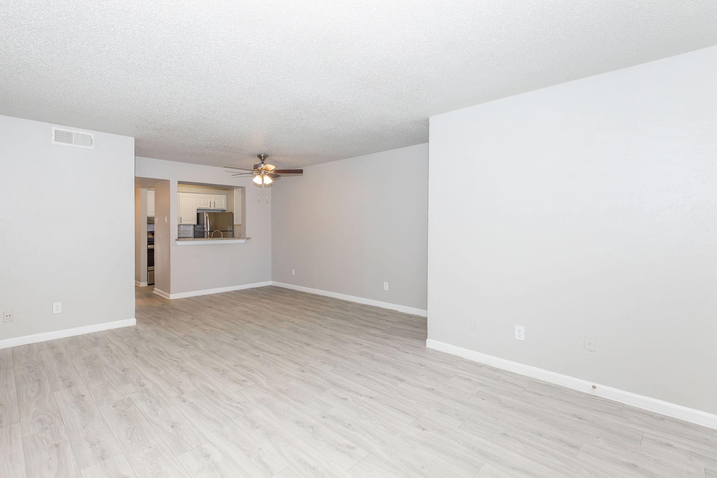 Empty living room with light gray walls and a ceiling fan. The floor features light-colored laminate. A doorway leads to a kitchen area in the background, visible through an open space in the wall. The room is well-lit, showcasing a spacious and modern design.