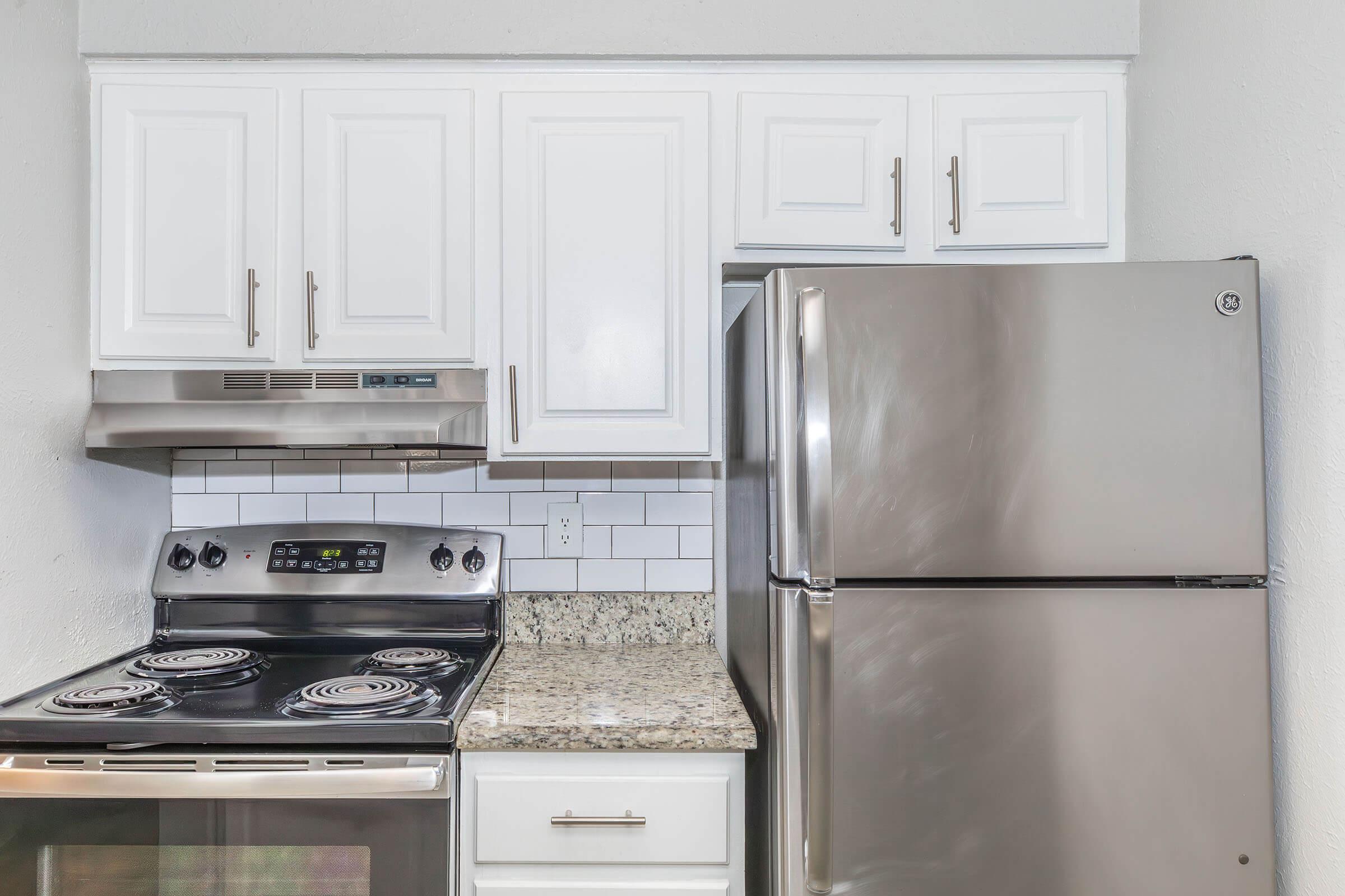 Modern kitchen setup featuring white cabinetry, a stainless steel refrigerator, a gray stove with a black cooktop, and a granite countertop. The backsplash is made of white subway tiles, creating a clean and contemporary look.