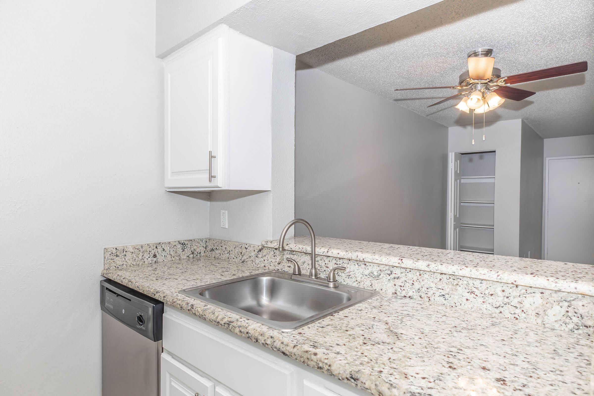 A modern kitchen with a granite countertop, stainless steel sink, and a dishwasher. The cabinetry is white, and there is a ceiling fan installed. The background shows an open space leading to a closet or storage area. The overall color scheme is light and neutral.