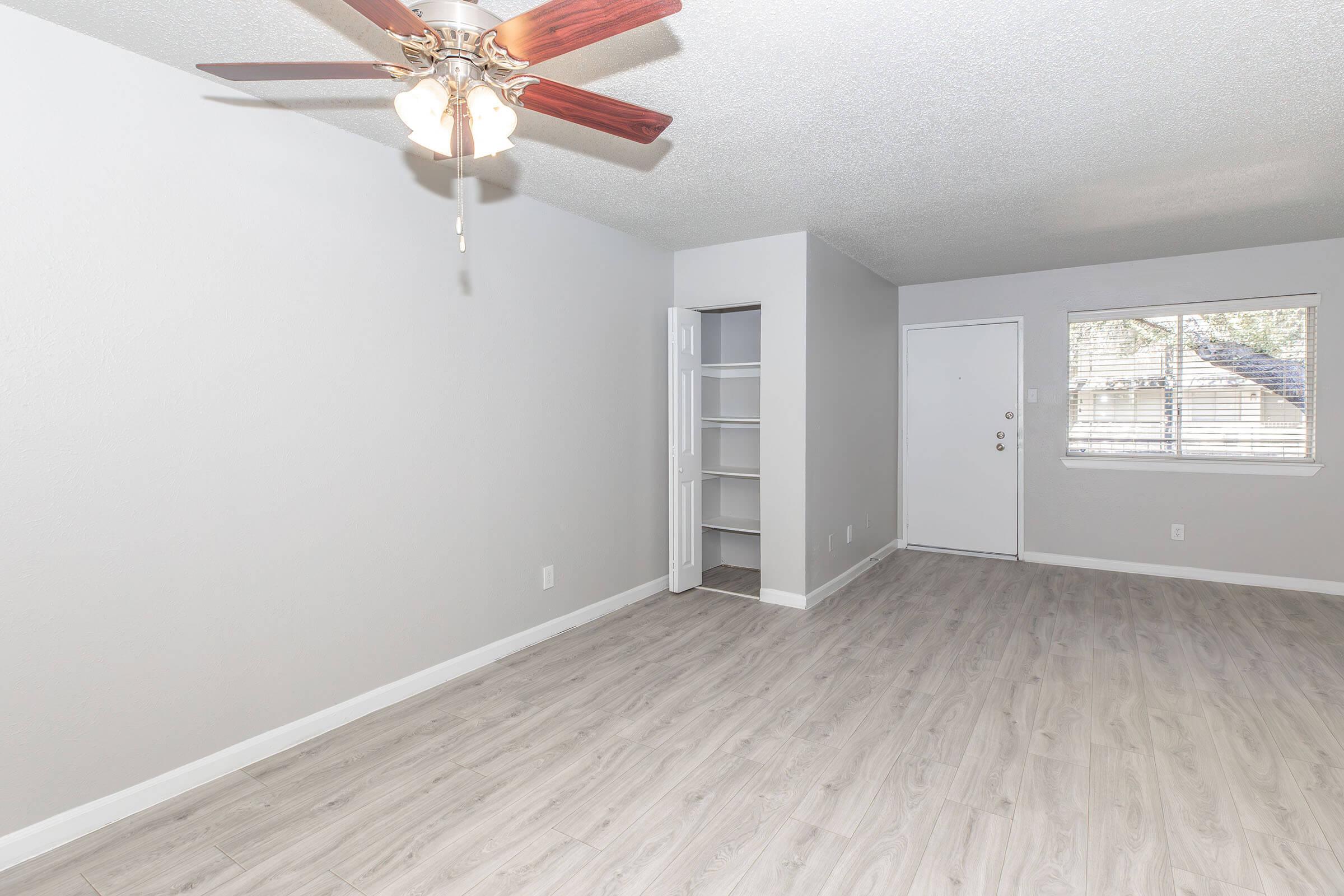 A spacious living room featuring light gray walls and wood-style flooring. There's a ceiling fan above, a small closet on the left, and a front door with a window next to it. Natural light streams in through the window, creating a bright and inviting atmosphere.