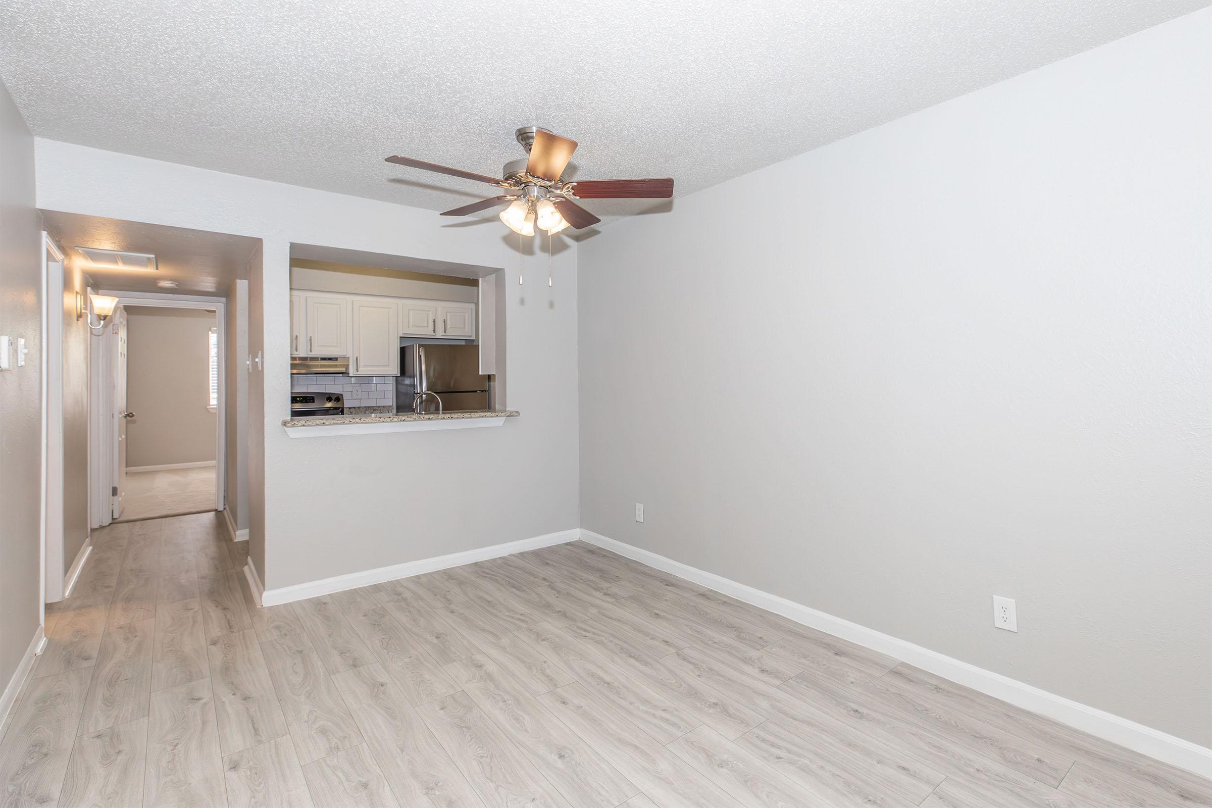 Interior view of a modern, empty living room featuring light-colored walls, wooden flooring, and a ceiling fan. An open layout connects to a kitchen area with white cabinetry in the background. There is a hallway leading to another room visible on the left.