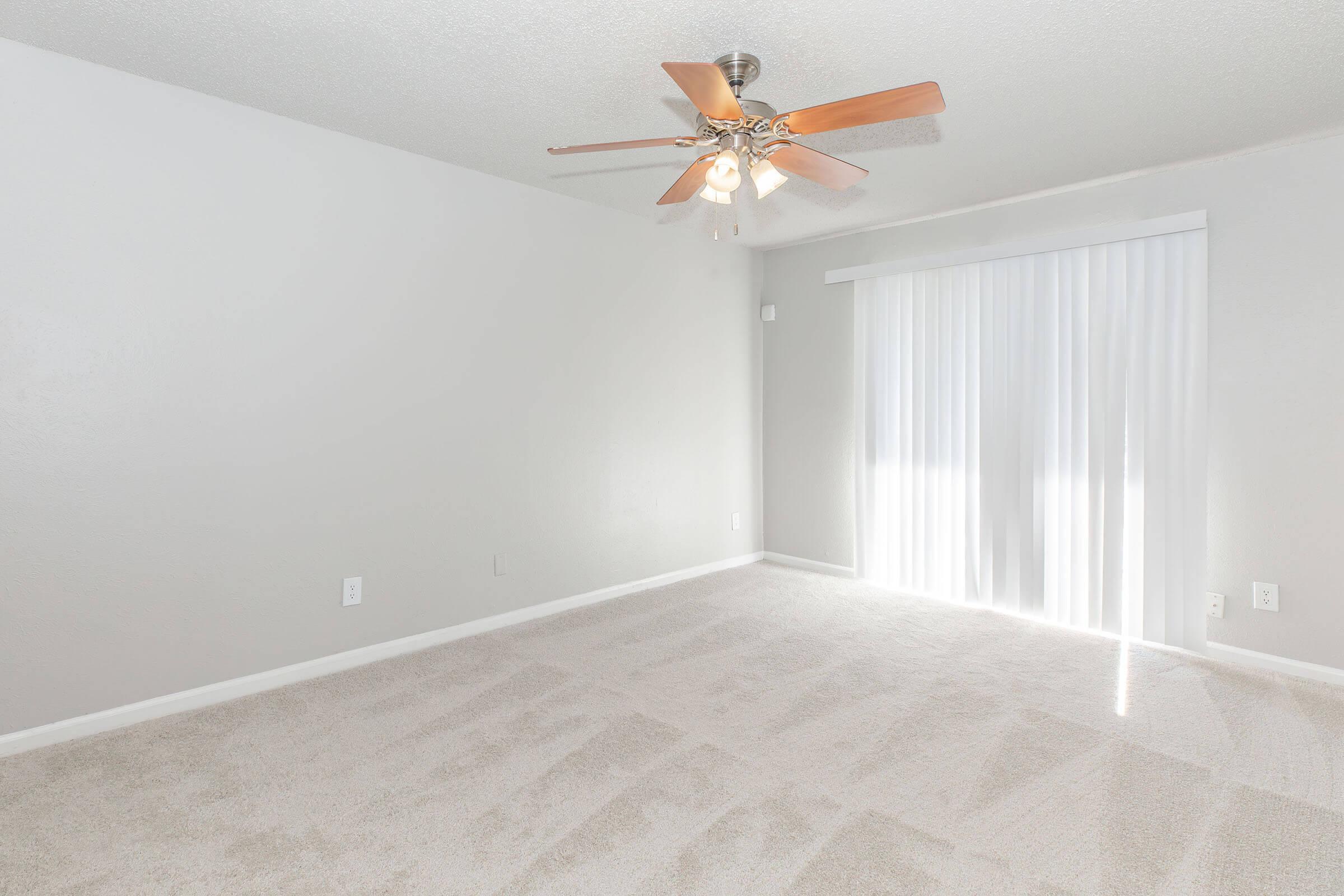 Bright and spacious room featuring light gray walls and plush carpet. A ceiling fan with wooden blades is installed, and a large window covered with vertical blinds allows natural light to enter the space. The overall ambiance is clean and inviting, ideal for various uses such as a living area or bedroom.