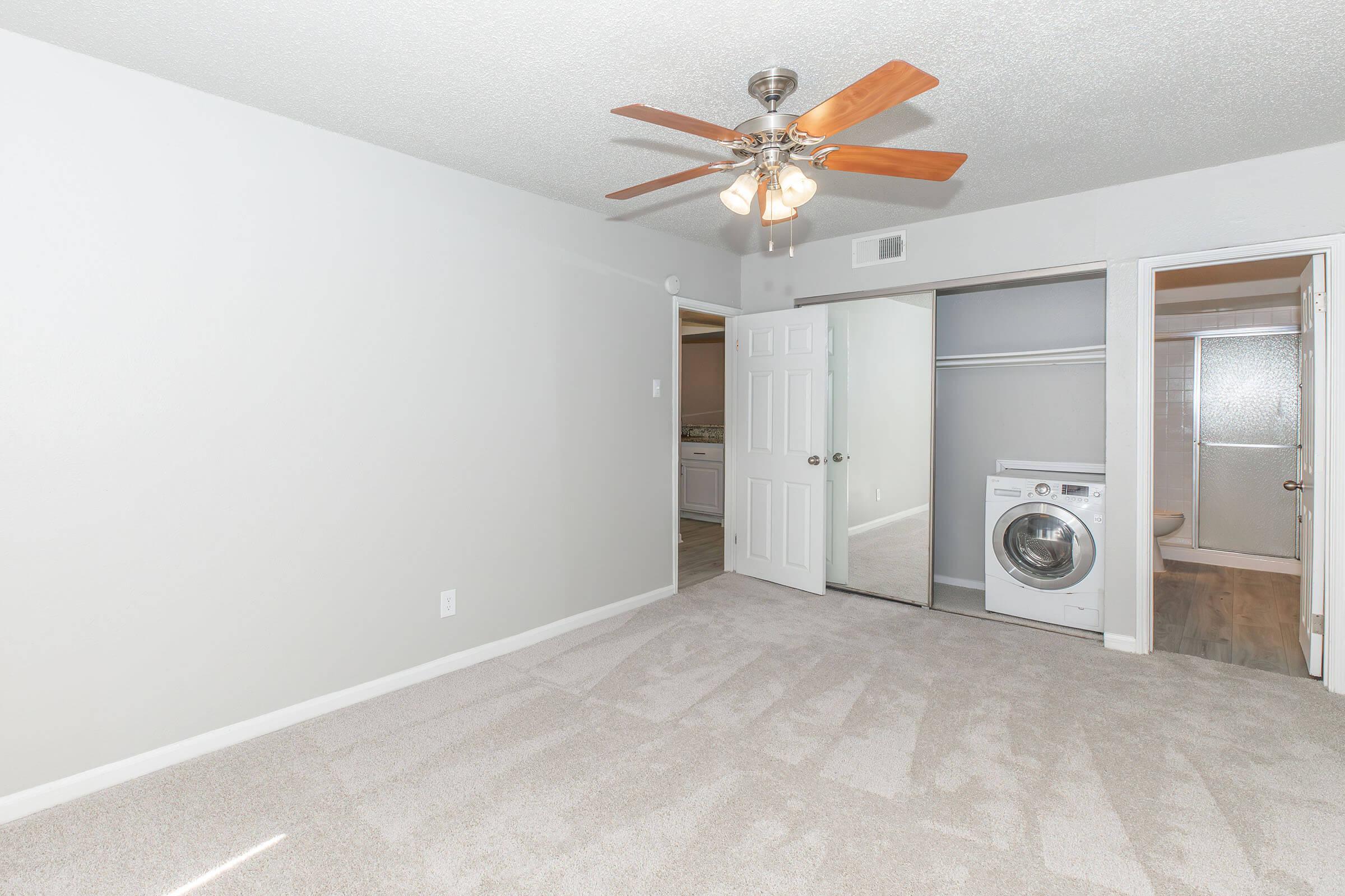 A spacious bedroom featuring light gray walls and a ceiling fan with wooden blades. There is a large window for natural light. To the right, a door leads to a bathroom, and there's a closet with a laundry machine visible inside. The floor is carpeted in a neutral tone.