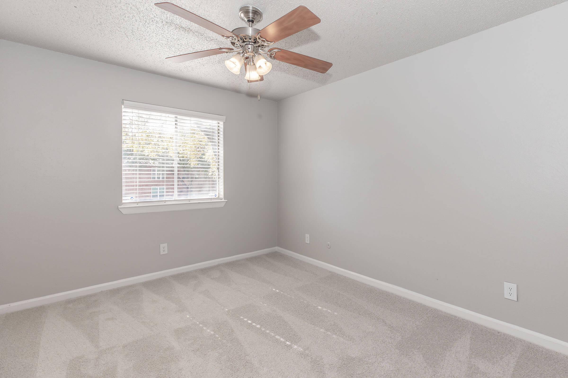 A well-lit, empty room with light gray walls and a ceiling fan. It has a window with white blinds allowing natural light in. The floor is covered with clean, beige carpet, and there are no furnishings or decorations present, providing a simple and spacious feel.