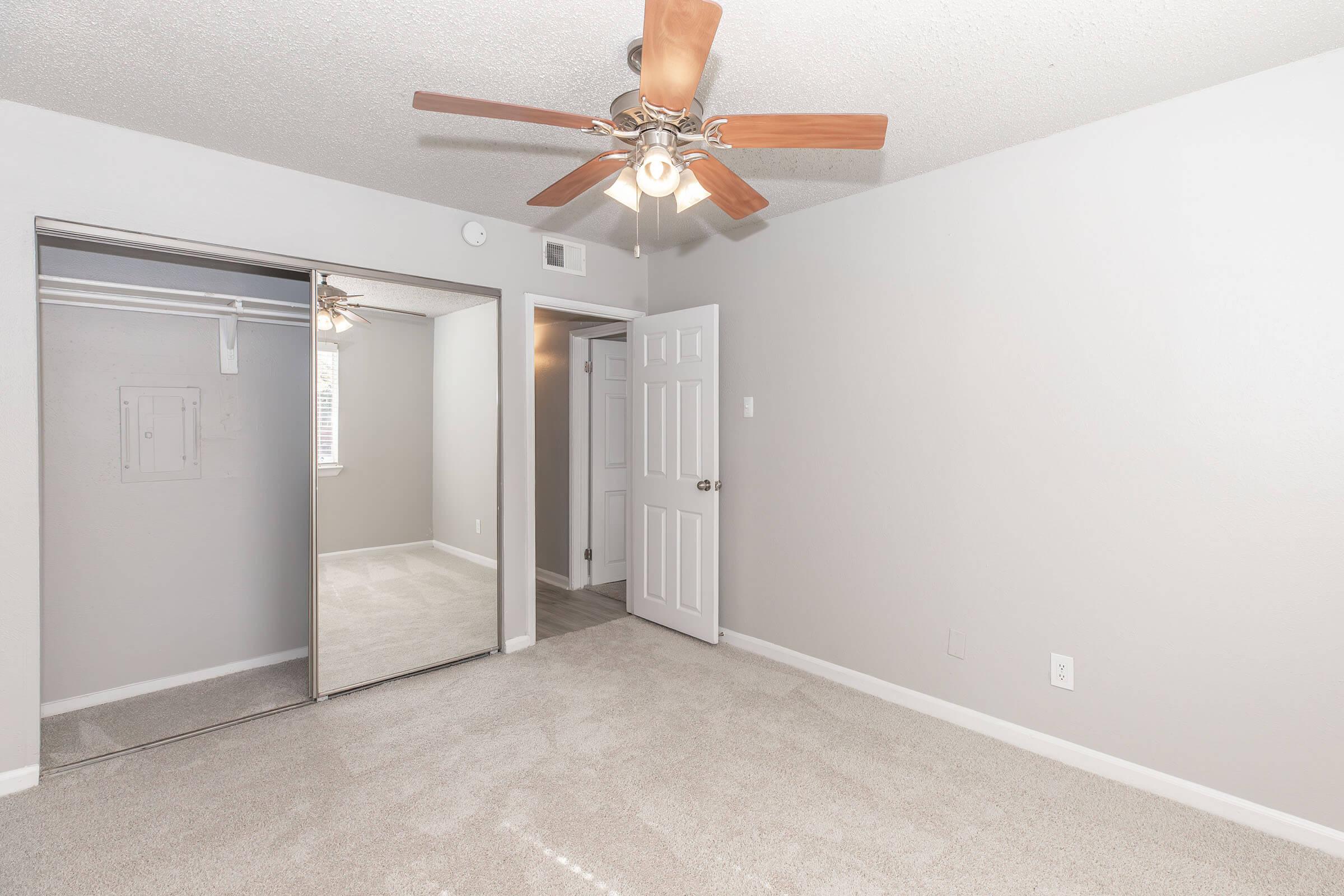 A spacious room with light gray walls, plush beige carpet, and a ceiling fan with wooden blades. Features a mirrored closet door and a white door leading to another room. Natural light enters through a window, creating an inviting atmosphere.