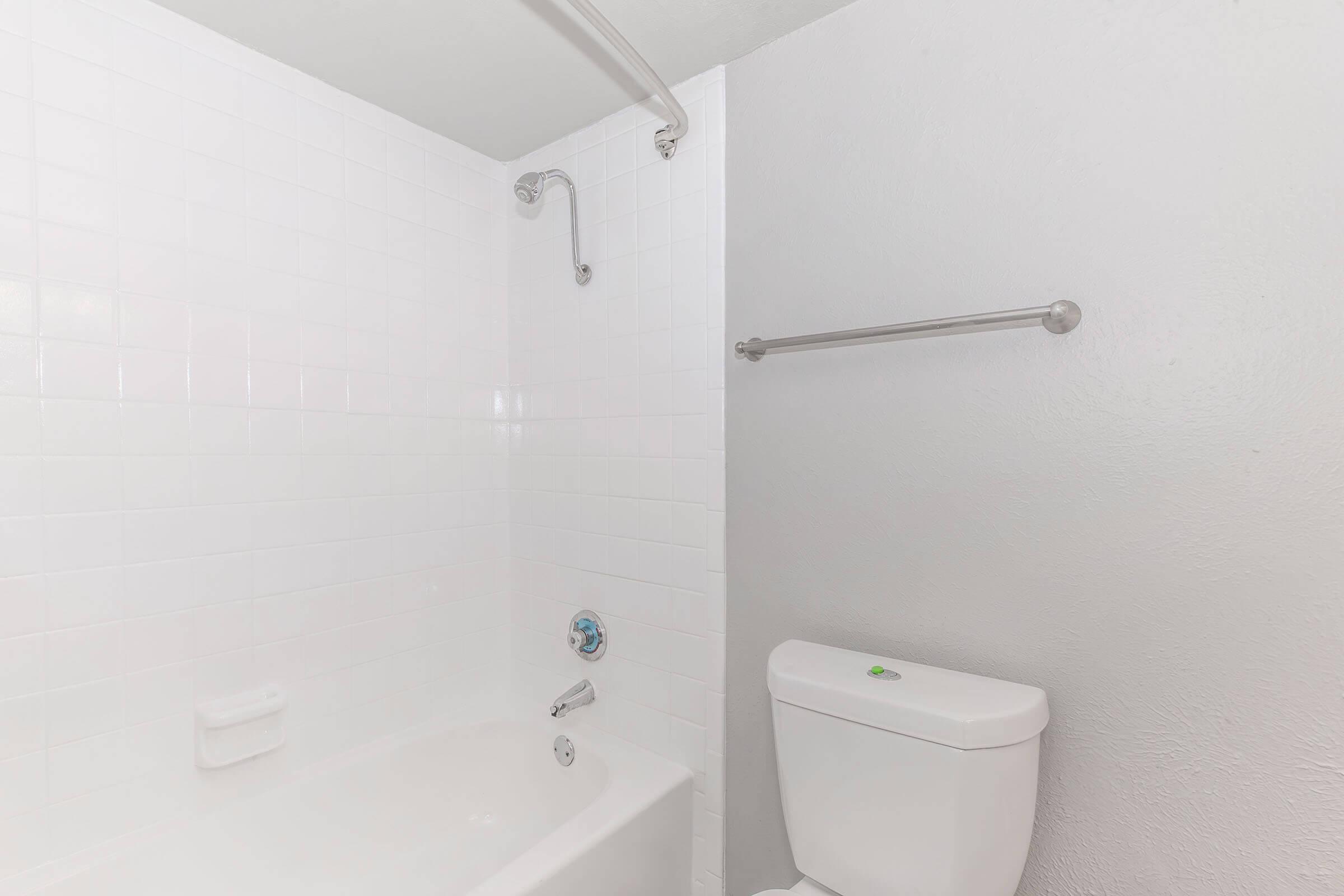 A clean, minimalistic bathroom featuring a white tiled bathtub with a shower overhead, a toilet next to it, and a grab bar on the wall. The walls are painted white, enhancing the bright and spacious appearance of the room.