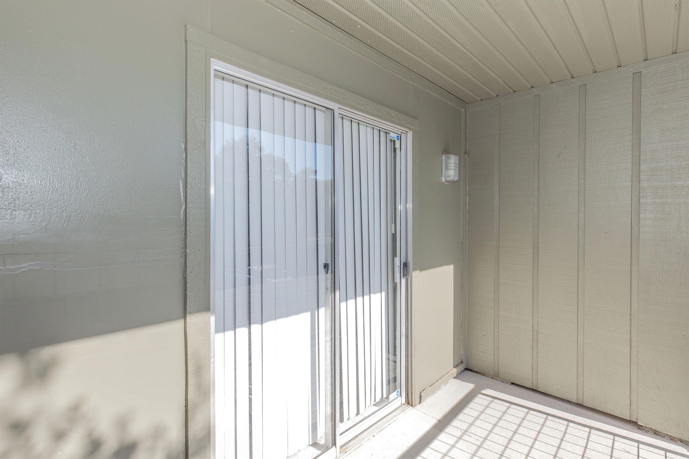 A corner view of a patio with sliding glass doors and vertical blinds. The walls are painted a light gray, and natural light filters in, casting shadows on the floor. The space appears clean and uncluttered, ideal for outdoor access.